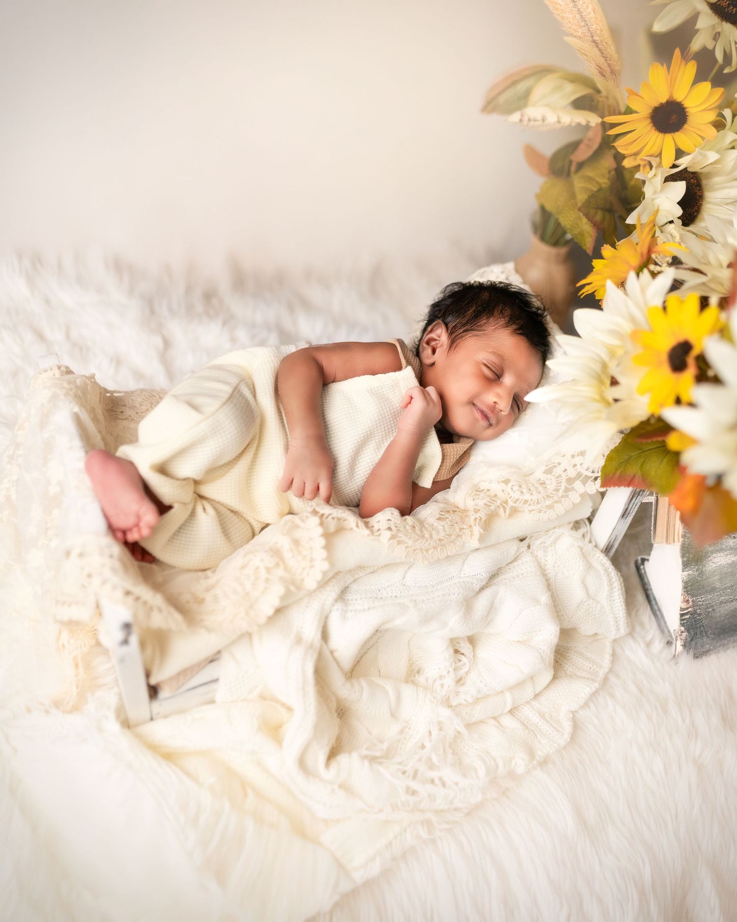 A sleeping baby in cream-colored clothing lies on white bedding with sunflowers in the background.
