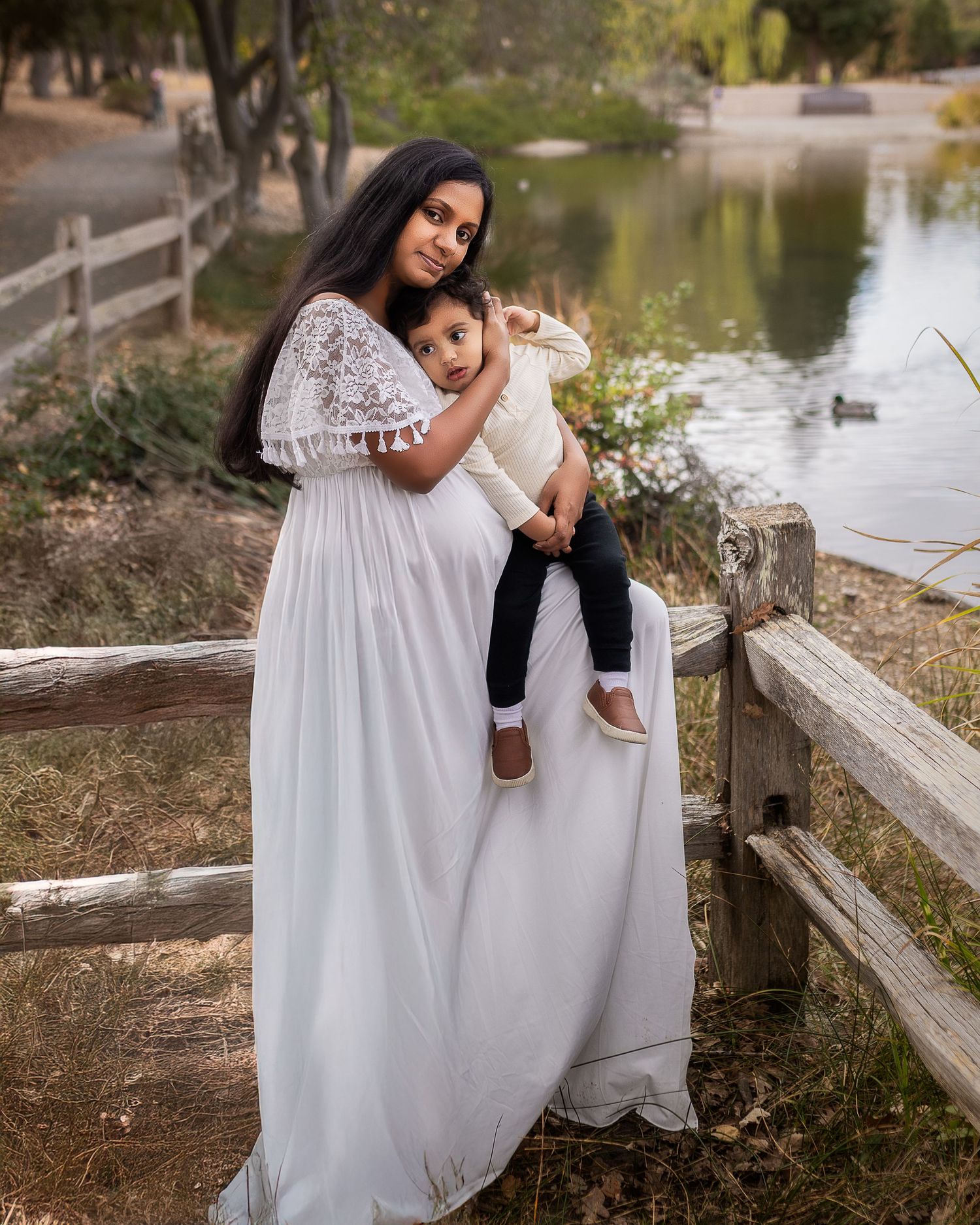 A white lace maternity gown worn in a serene park setting with a wooden fence and pond in the background.