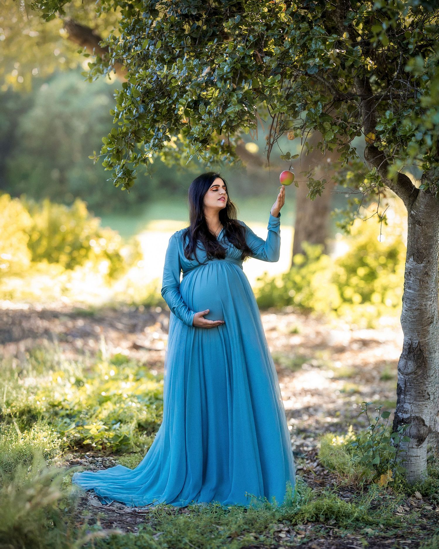 A figure in a flowing turquoise gown poses gracefully in a sun-dappled forest setting with trees and natural light.
