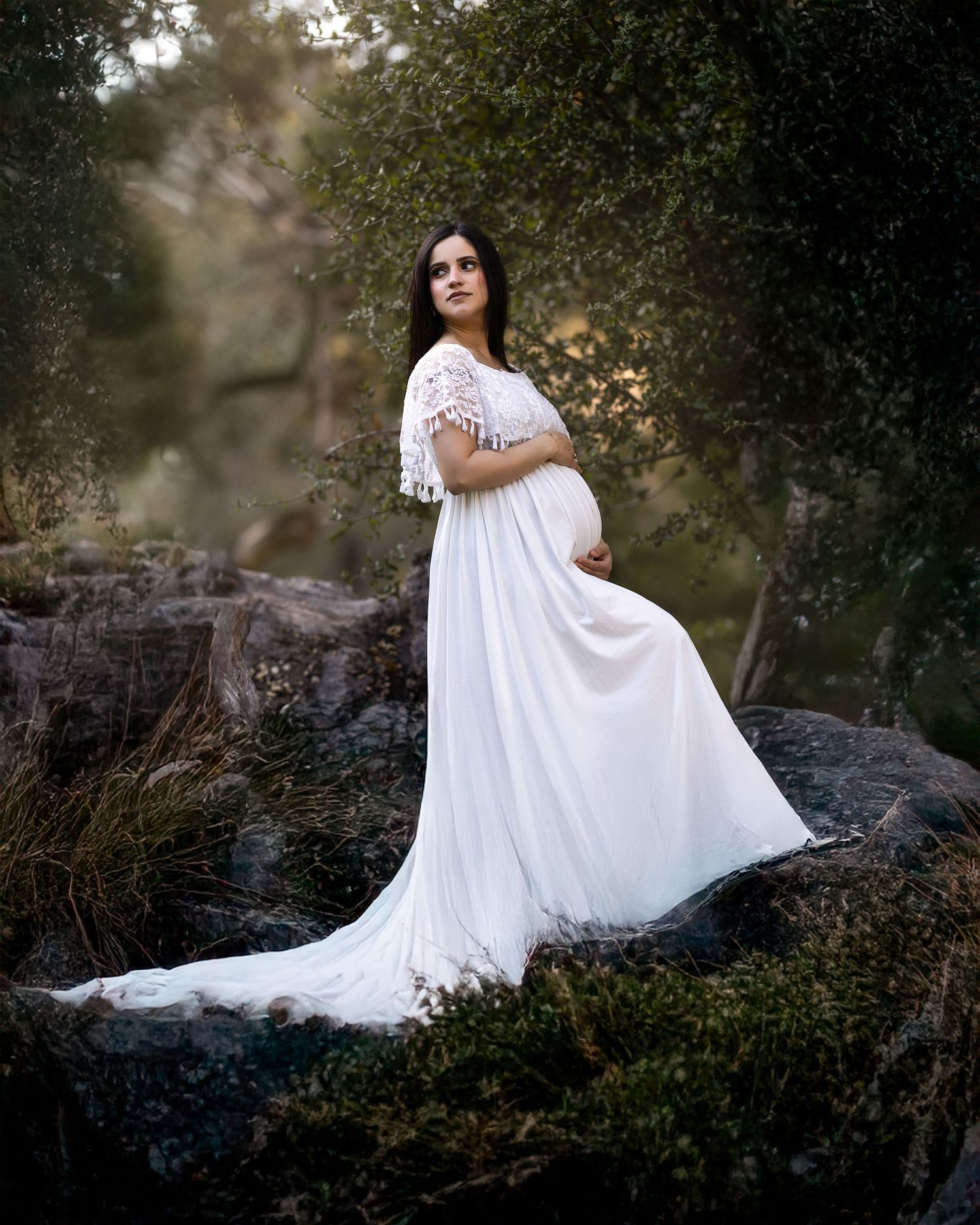 A flowing white lace dress creates an ethereal scene against dark rocks and foliage in a dreamy outdoor photoshoot.
