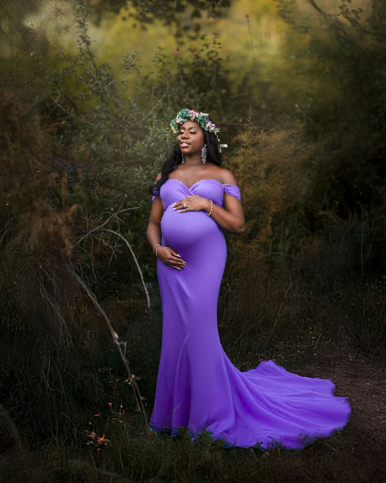 Expectant mother in flowing purple off-shoulder maternity gown and floral crown poses gracefully in dreamy outdoor setting.