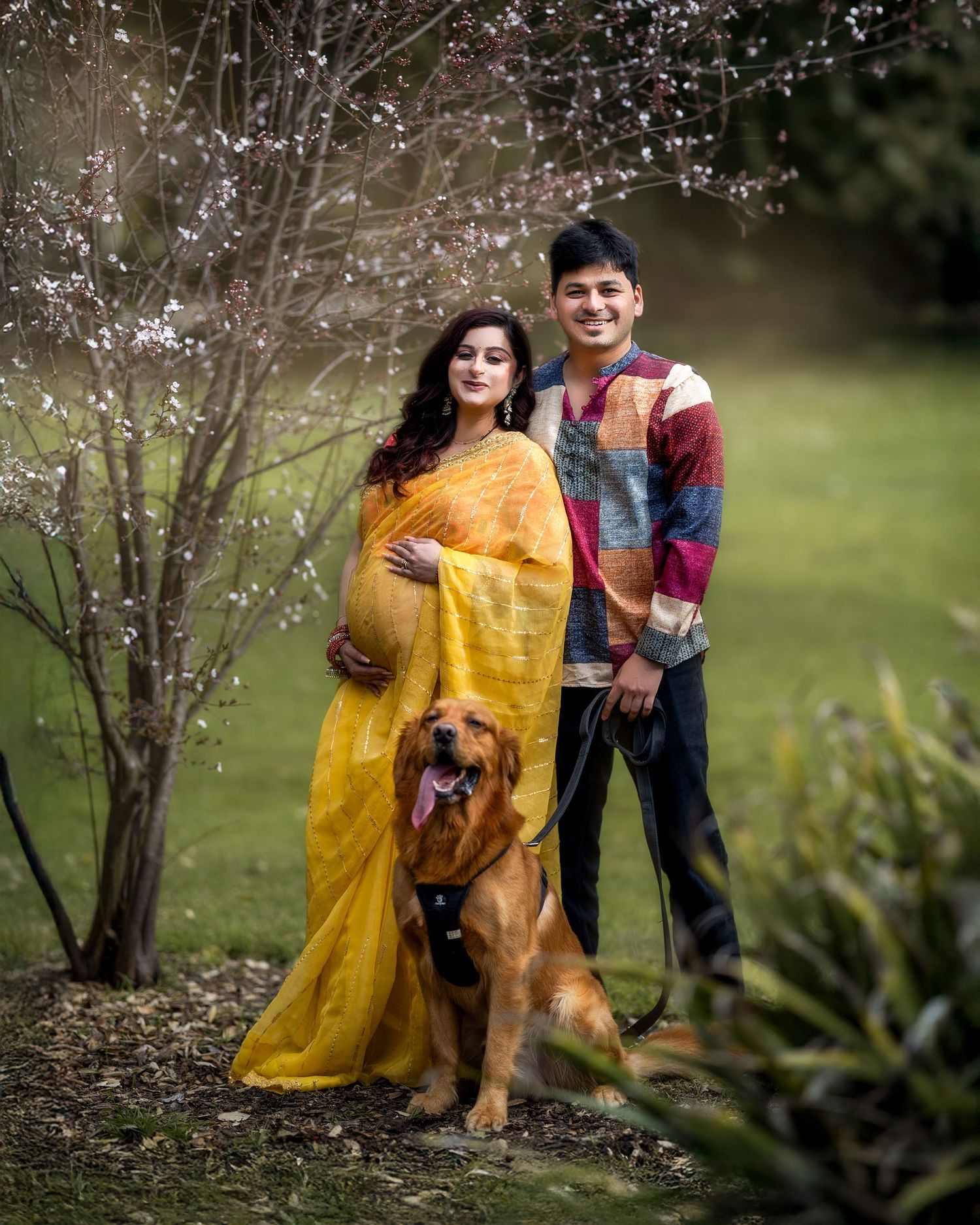 A family and their brown dog pose together in a garden setting with blooming trees and a bright yellow dress.