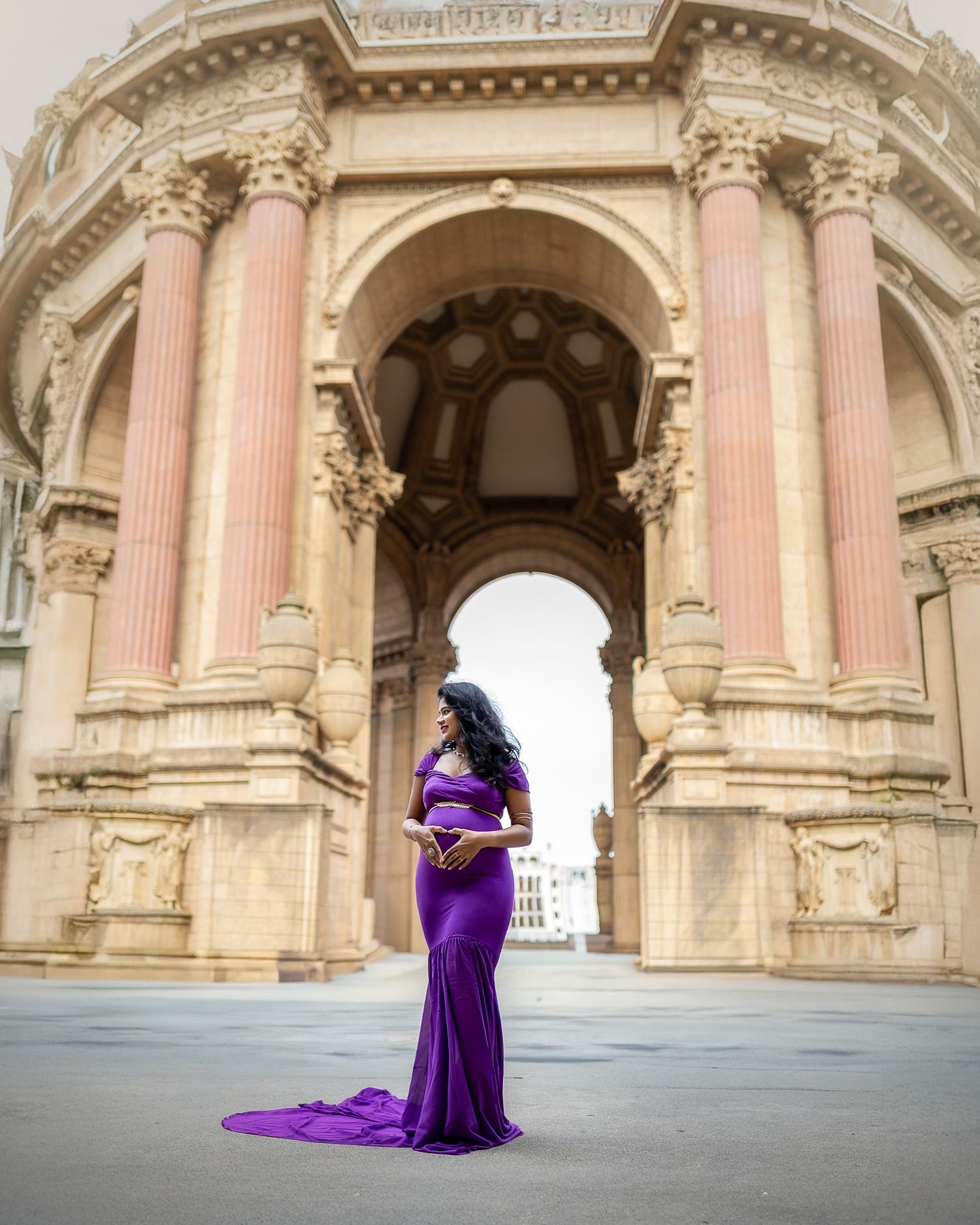 A person in a flowing purple gown poses gracefully under ornate classical stone archways with decorative columns.