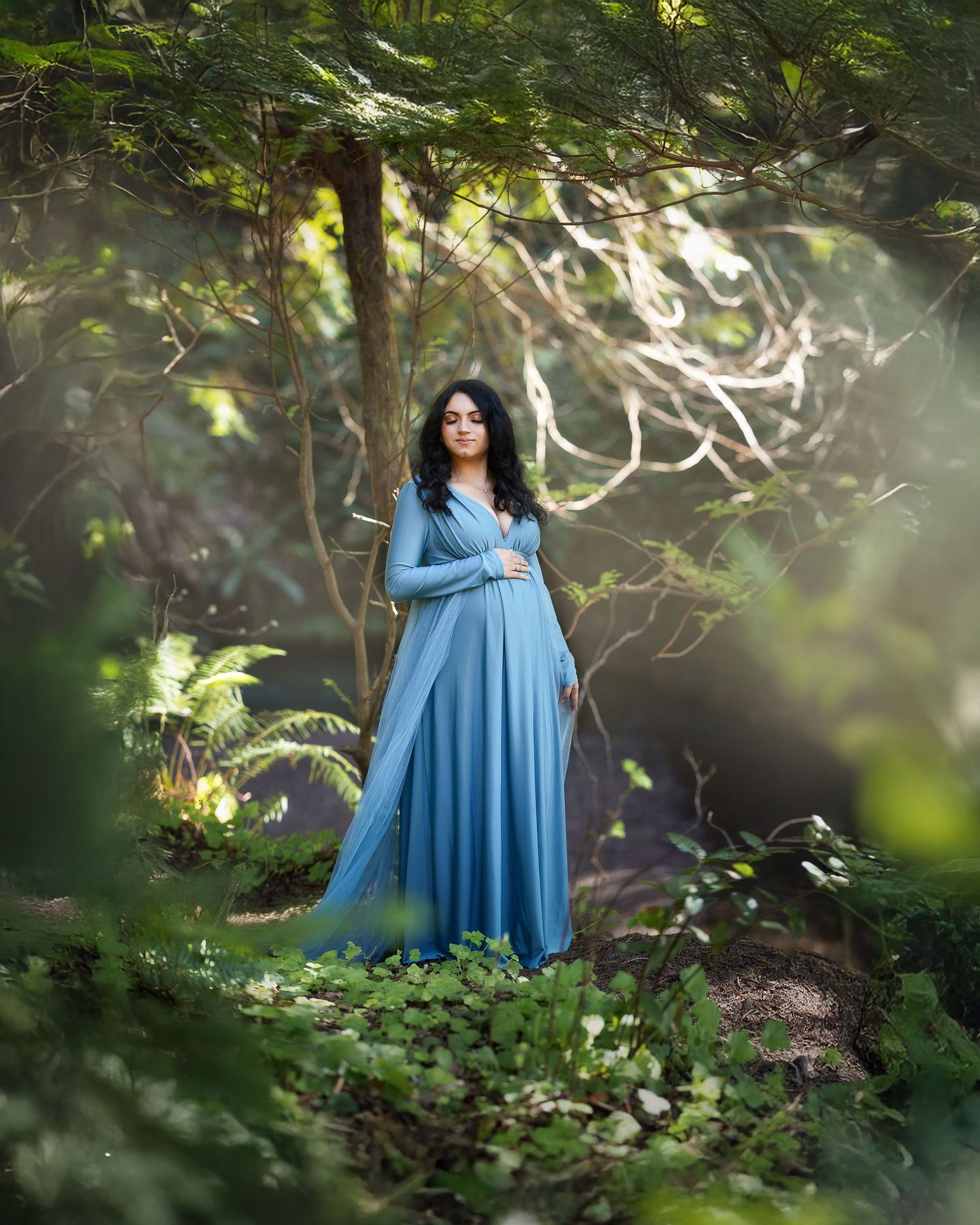 A dreamy maternity portrait in a blue flowing dress surrounded by lush forest greenery and dappled sunlight.