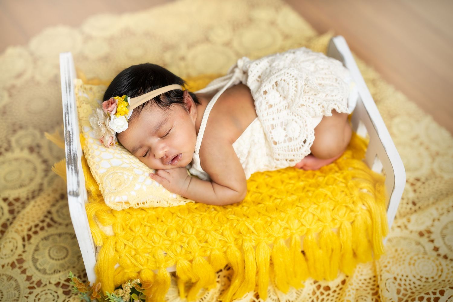 Sleeping newborn baby wrapped in yellow blanket with white lace trim rests on textured beige backdrop.