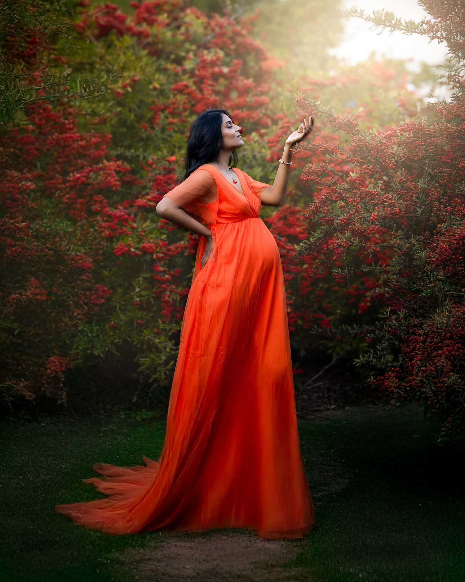 A figure in a vibrant orange gown dances against a backdrop of red flowering bushes in golden sunlight.