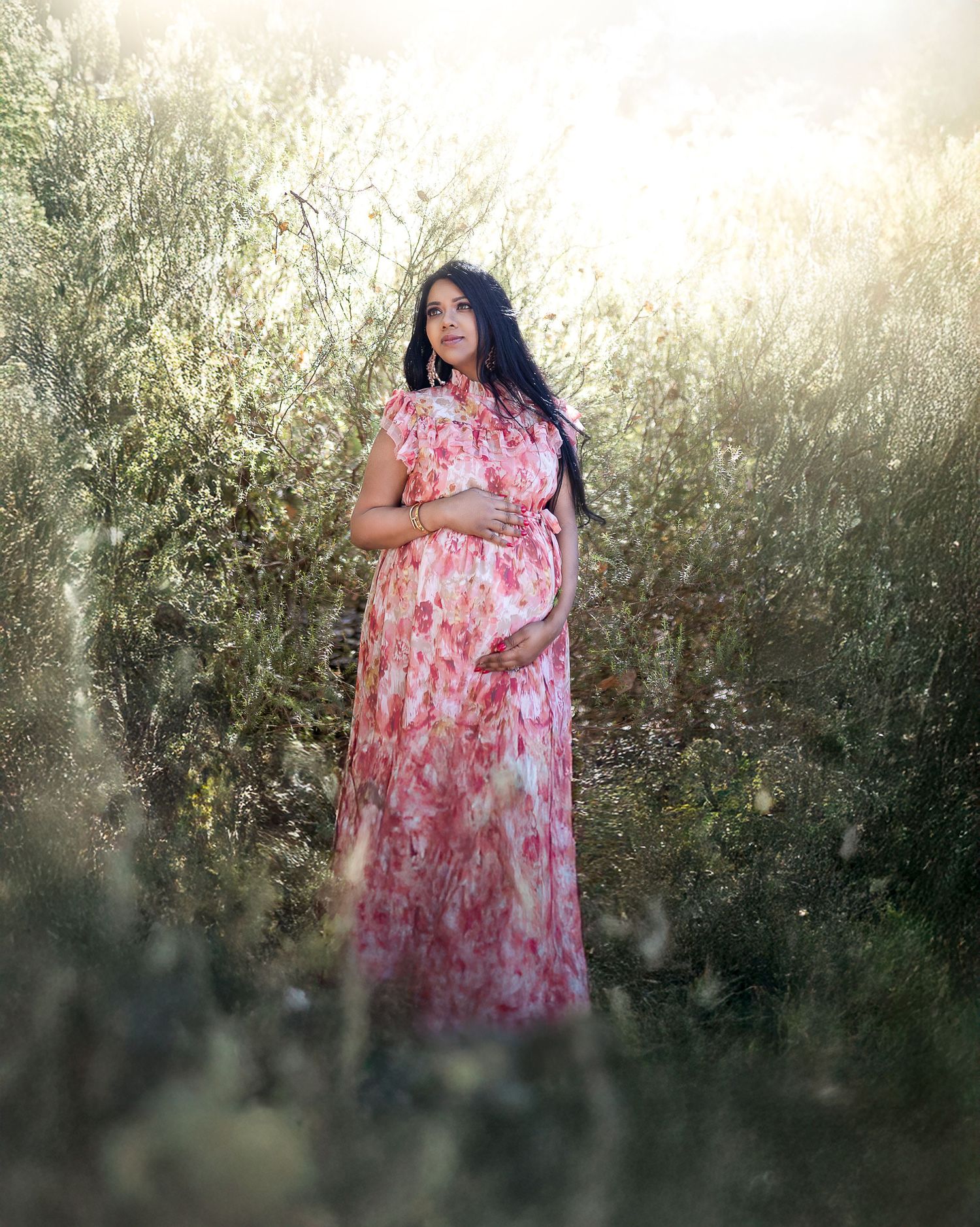 A person in a pink floral maternity dress stands in a sunlit garden with glowing backlight creating a dreamy atmosphere.