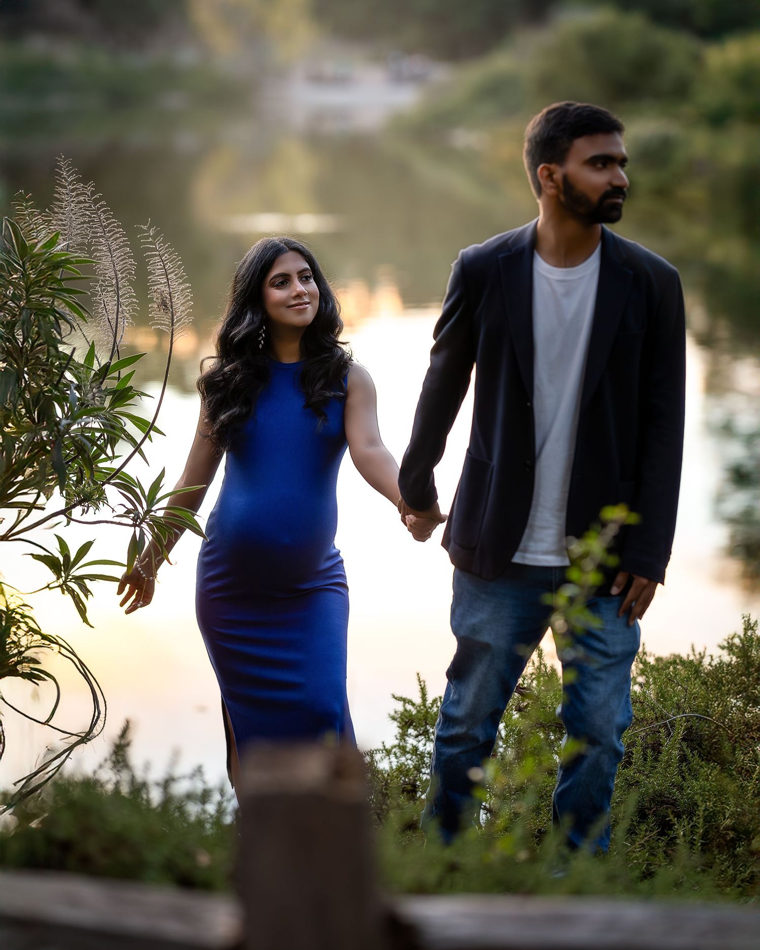 A couple holds hands while walking along a waterfront path at sunset, with the woman wearing a blue dress.