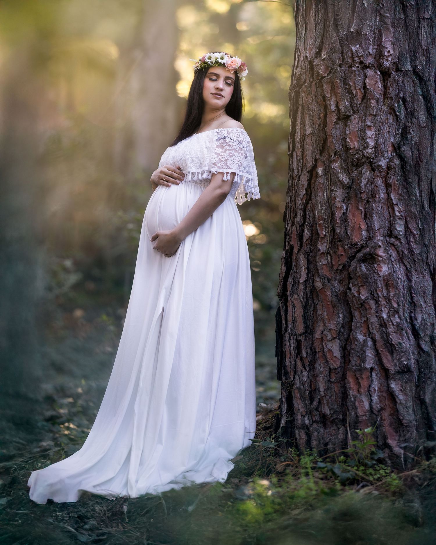 A dreamy maternity photo in a forest shows an ethereal white lace gown and floral crown against tall pine trees.