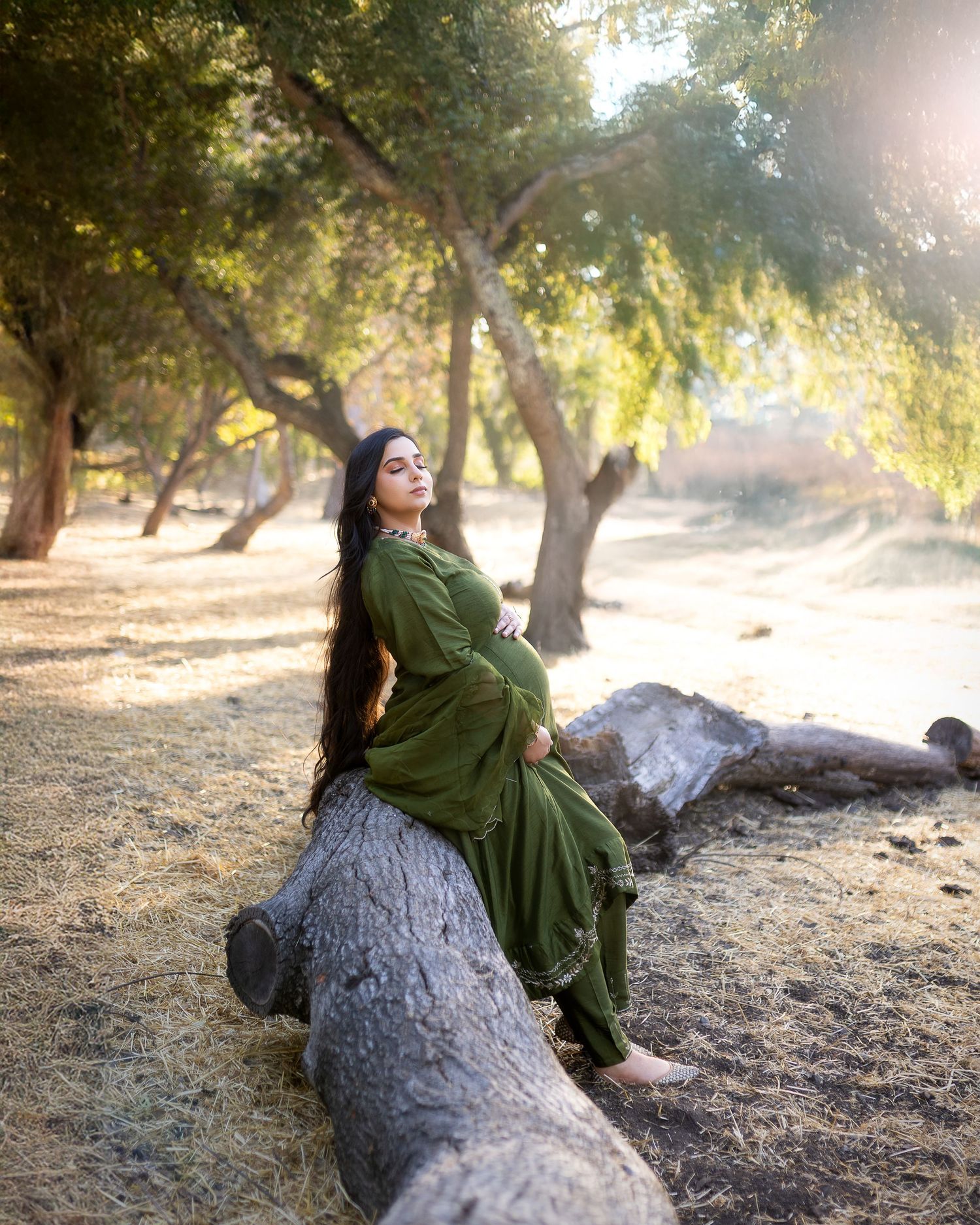 A figure in a flowing green dress poses artistically on a fallen tree trunk in a sunlit orchard setting.