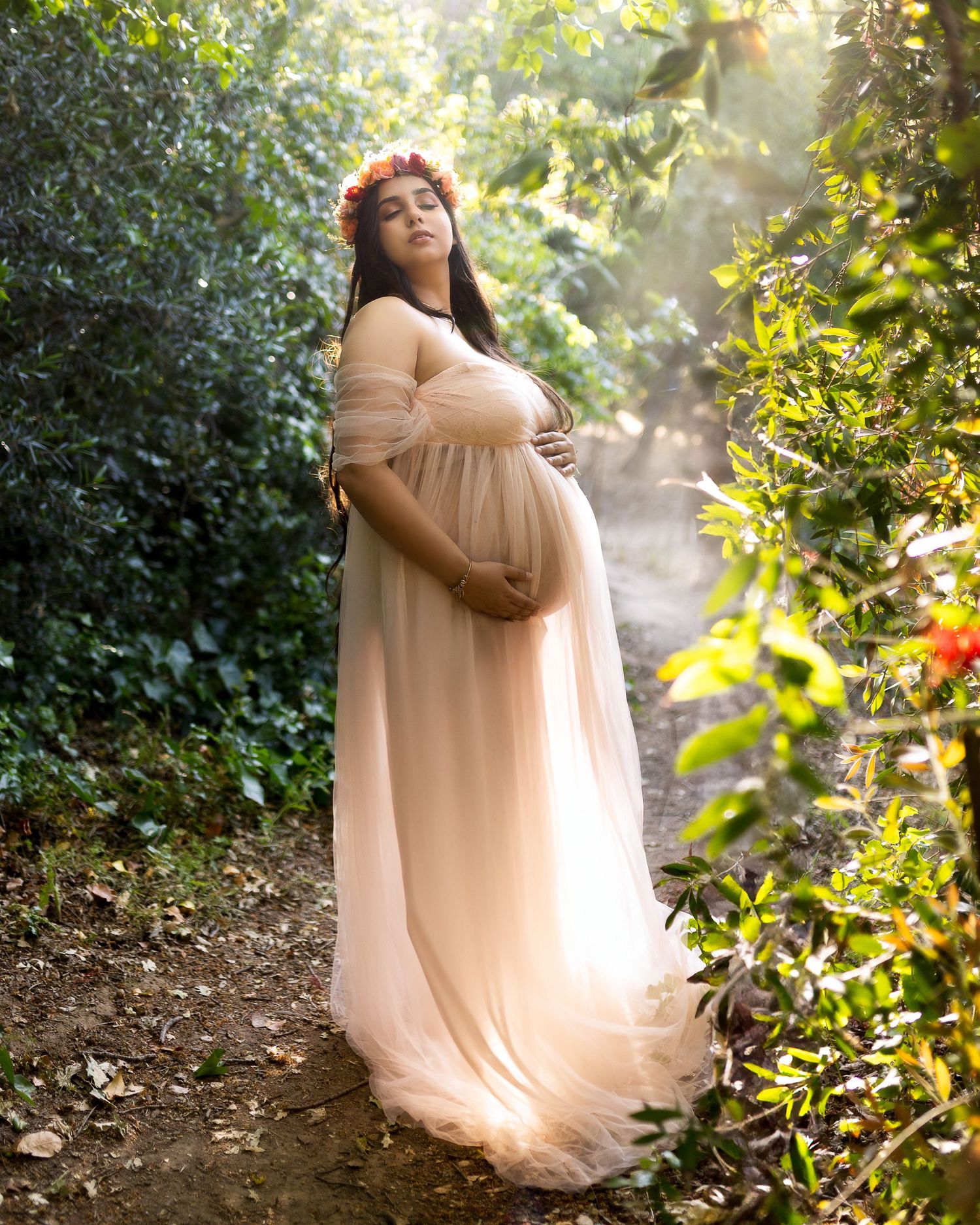 A figure in a flowing peach gown poses gracefully among sunlit trees and foliage in an outdoor maternity photoshoot.