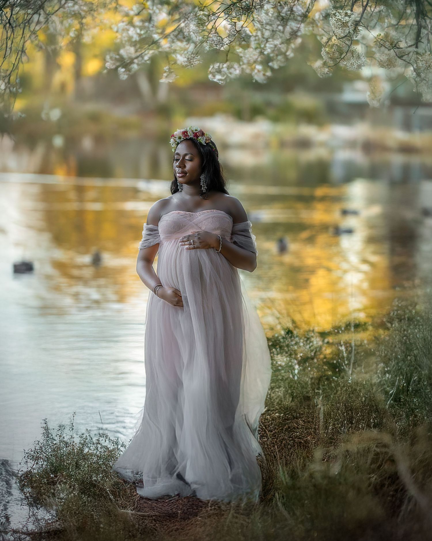 A pregnant woman in a flowing white dress stands by a lake with cherry blossoms during sunset.