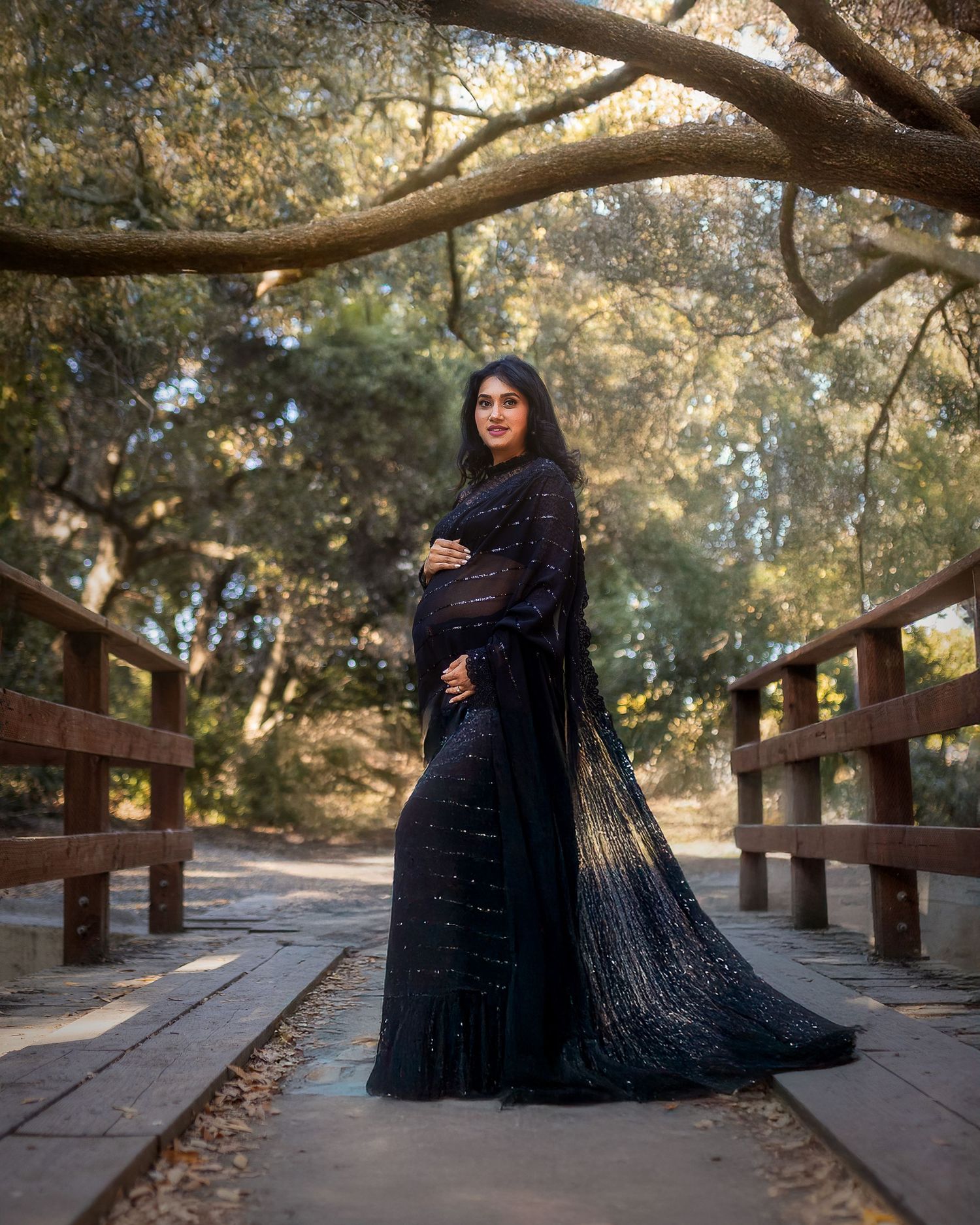 A figure in a flowing black gown poses dramatically on a wooden bridge surrounded by sunlit trees in a park setting.
