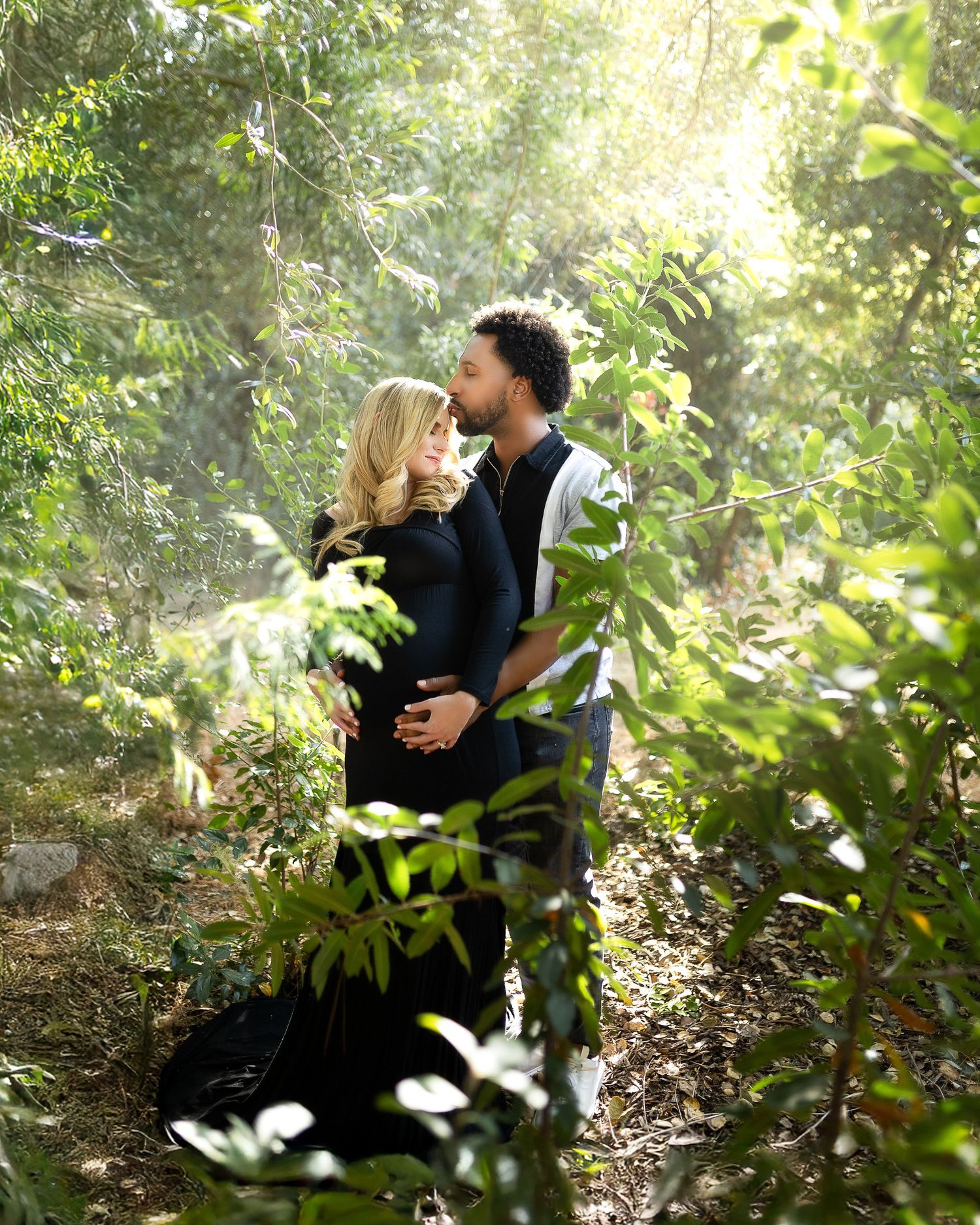 A romantic moment captured in a sun-dappled forest setting with soft natural lighting filtering through the trees.
