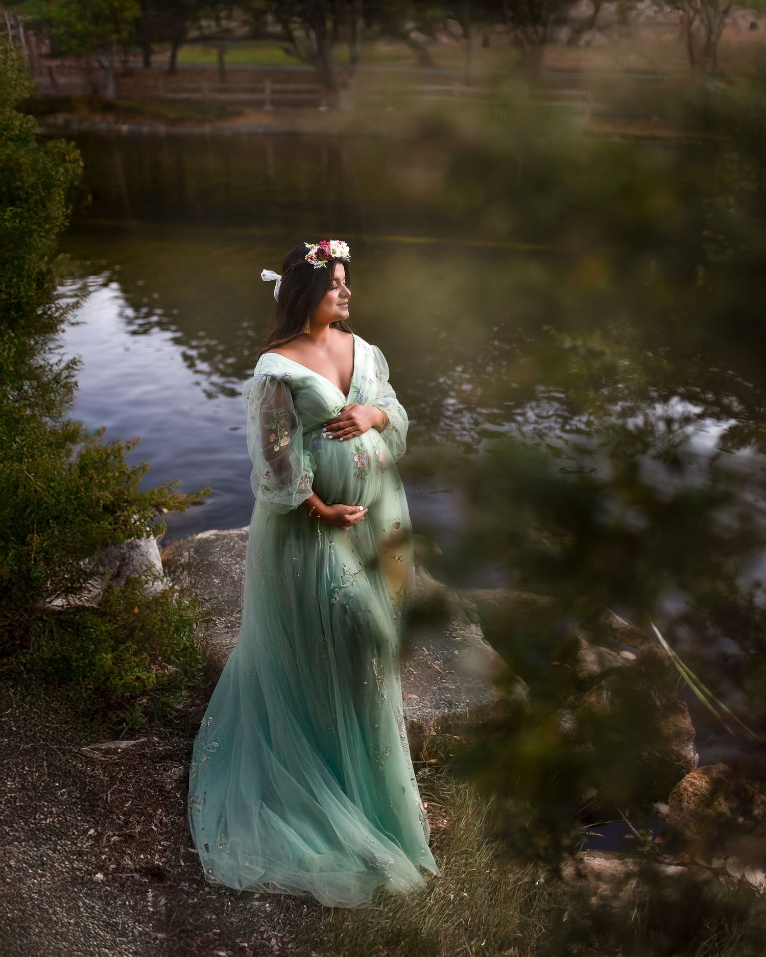 A dreamy maternity photo of a woman in a flowing mint green dress standing beside a serene pond.