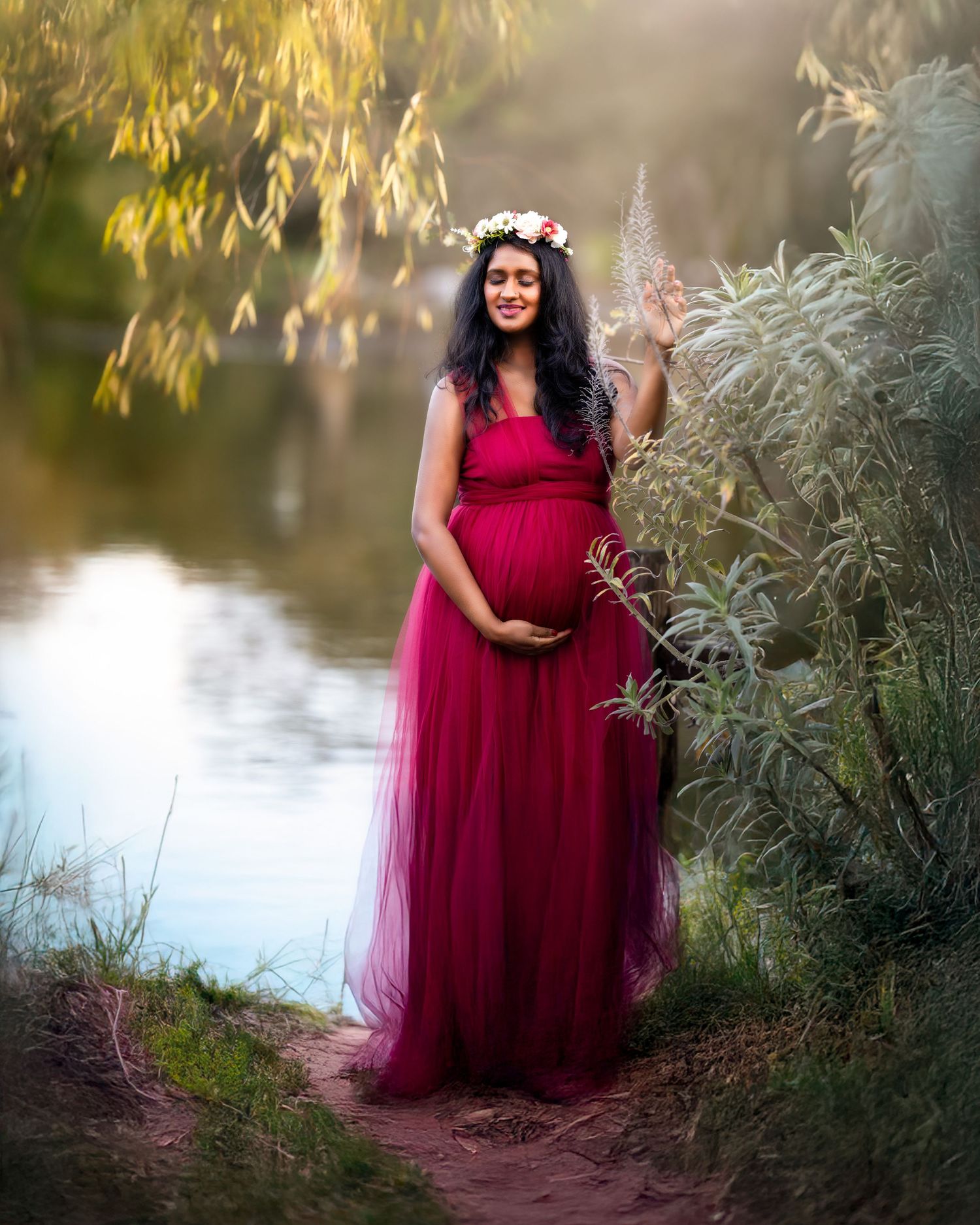 A pregnant woman in a flowing burgundy gown and floral crown poses gracefully by a tranquil pond at sunset.
