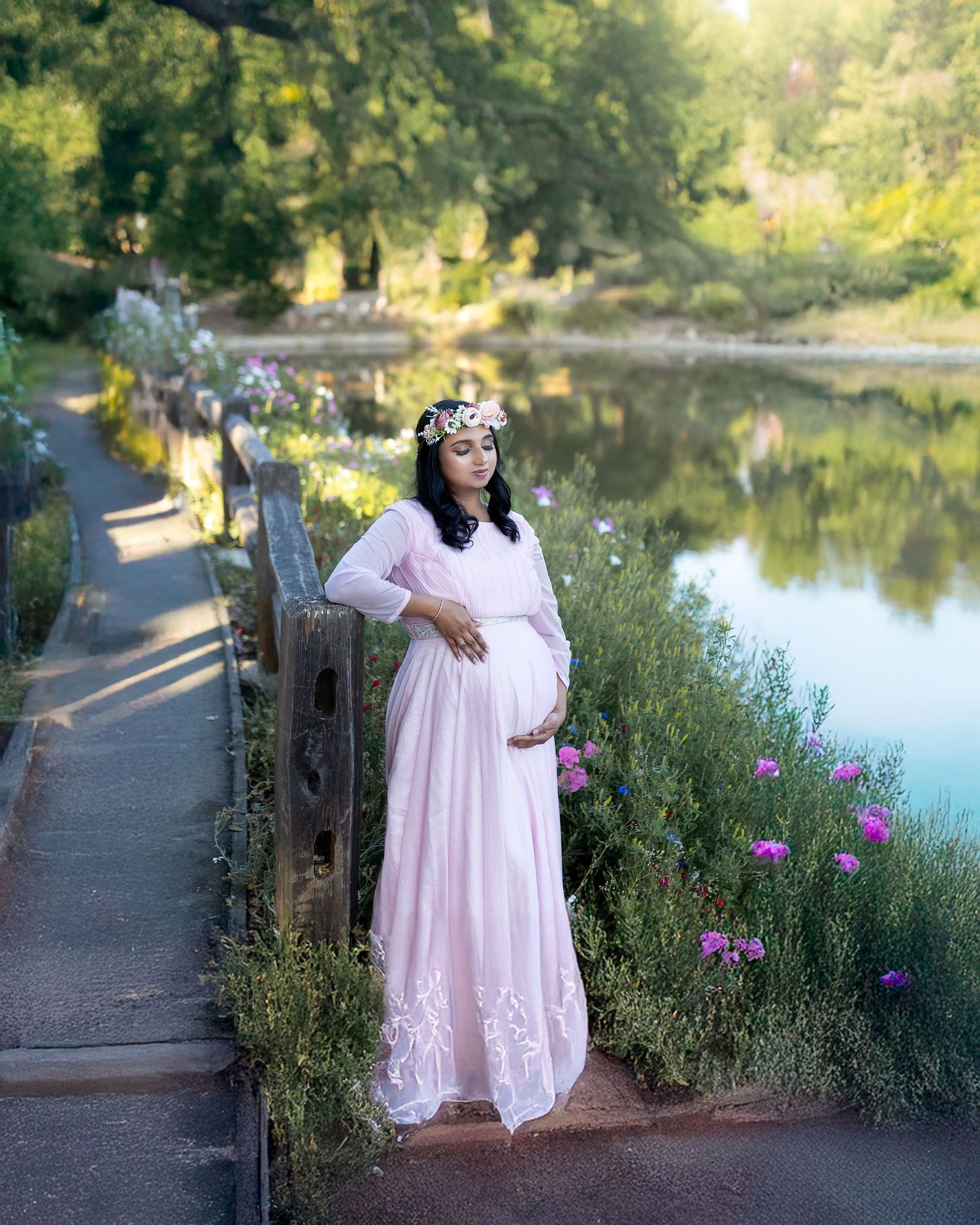 A person in a flowing pink dress poses by a serene lake surrounded by blooming flowers and greenery at sunset.