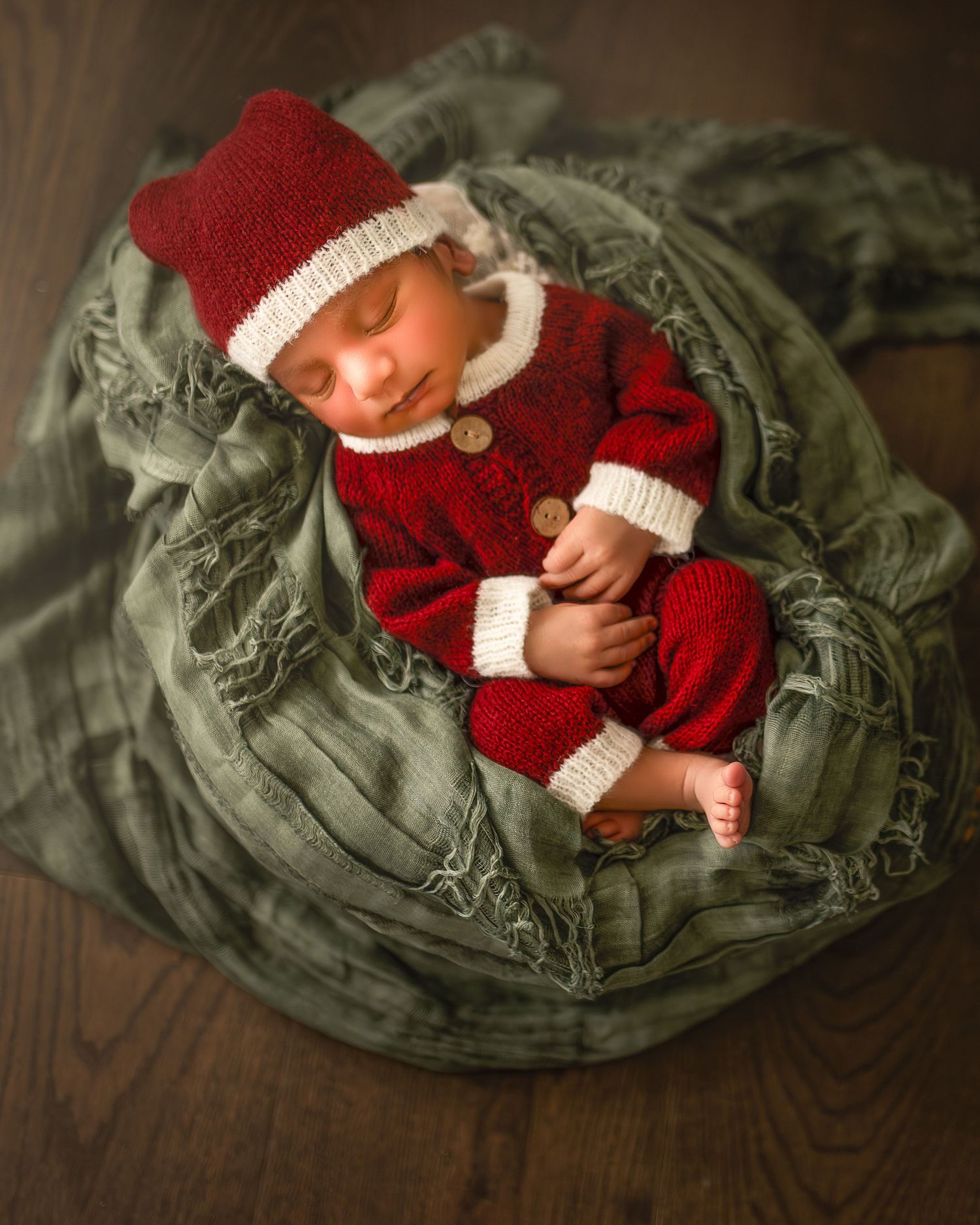 A sleeping infant wearing a festive red and white knit outfit rests on a sage green textured fabric backdrop.