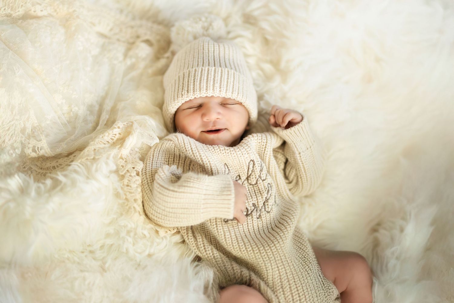 Sleeping baby wearing cozy cream knit sweater and beanie on soft white blanket.