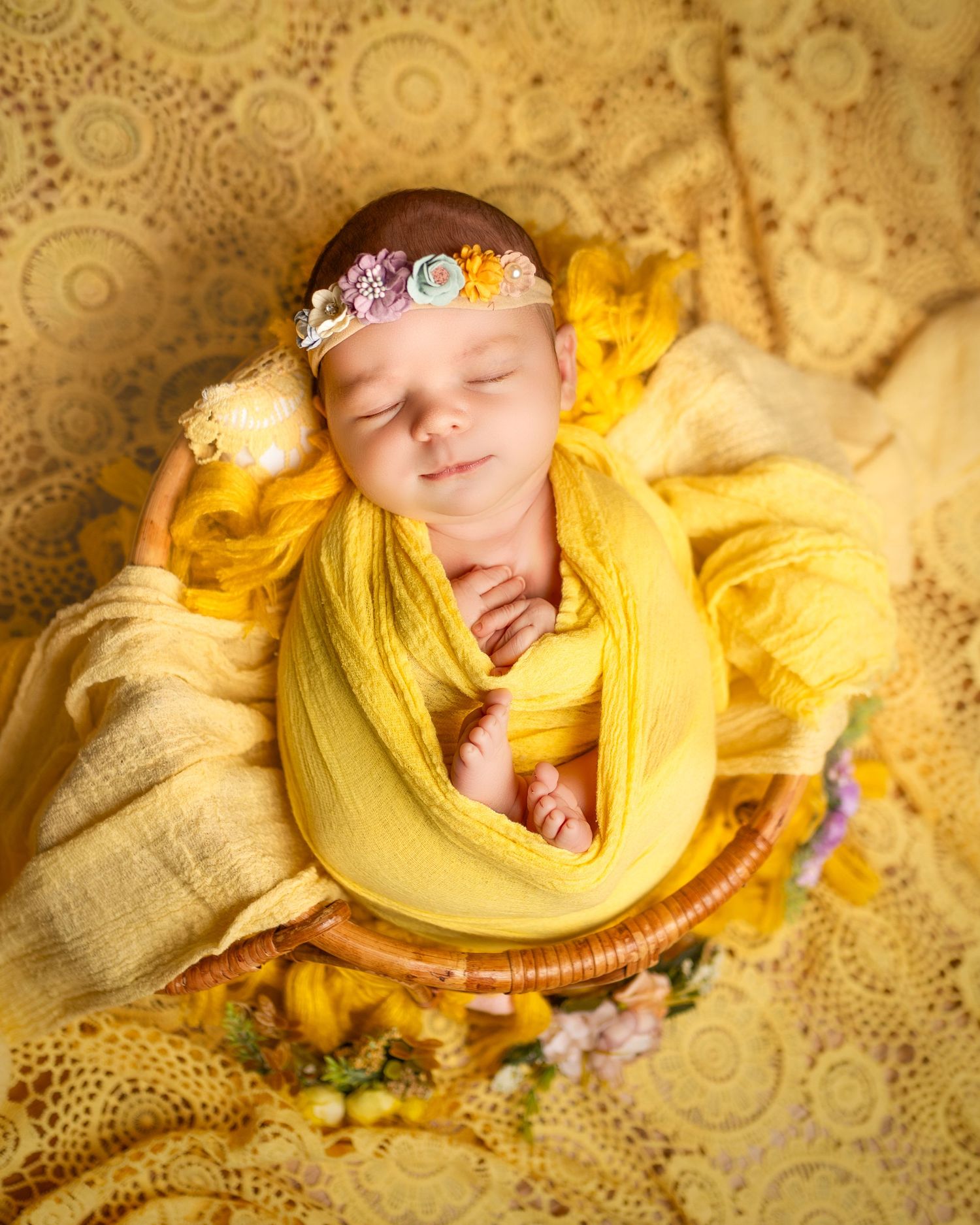 Sleeping newborn wrapped in soft yellow fabric on delicate lace blanket with purple flowers as decorative accents.