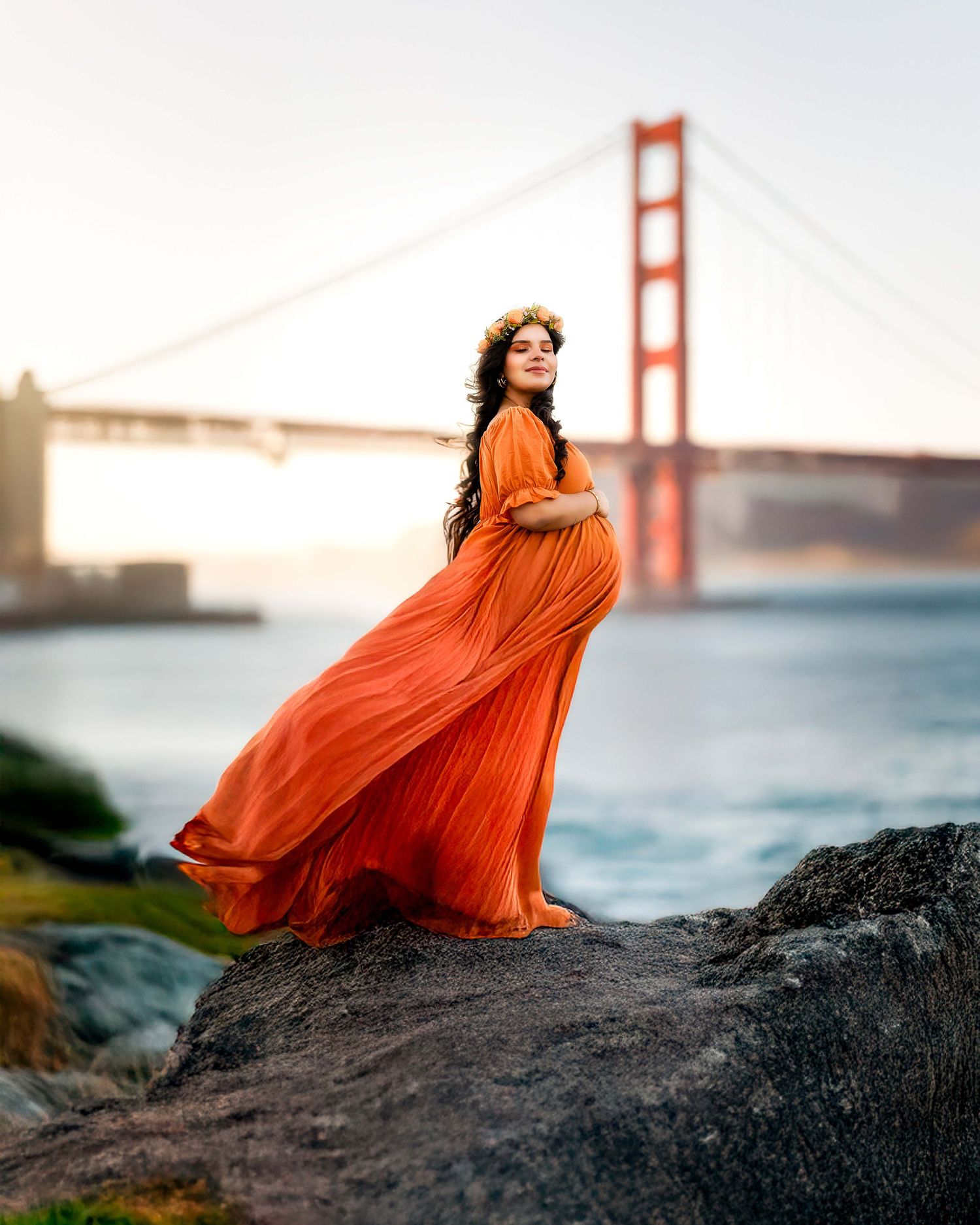 Pregnant woman in flowing orange dress stands on rocks with Golden Gate Bridge backdrop at sunset.