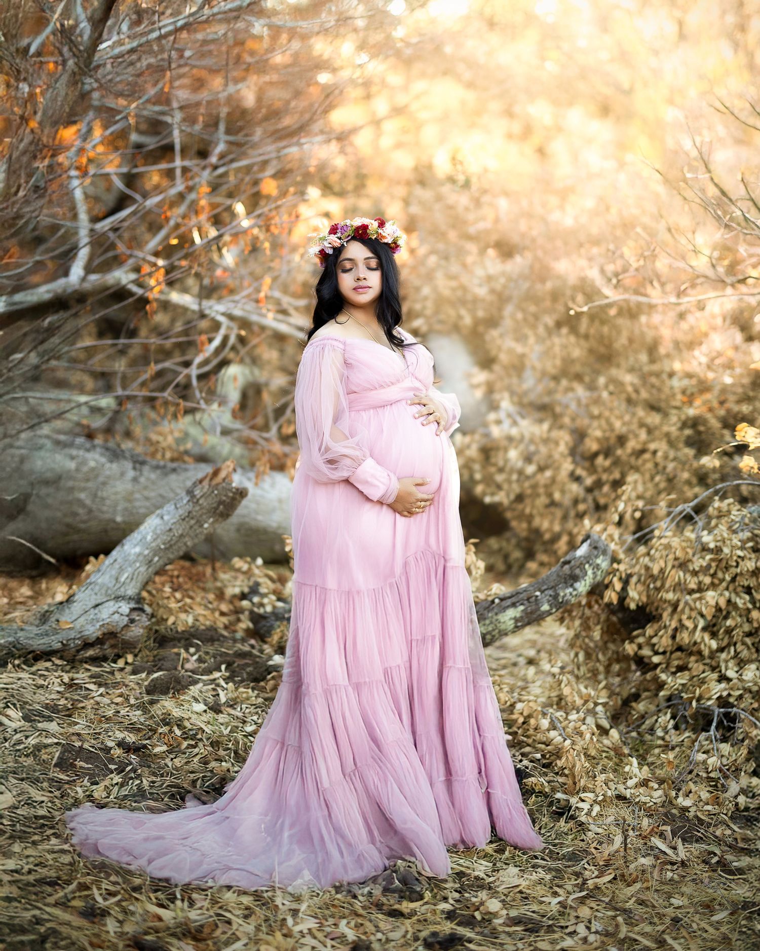 A maternity photo session in a forest with soft pink flowing dress and autumn colors creating a dreamy atmosphere.