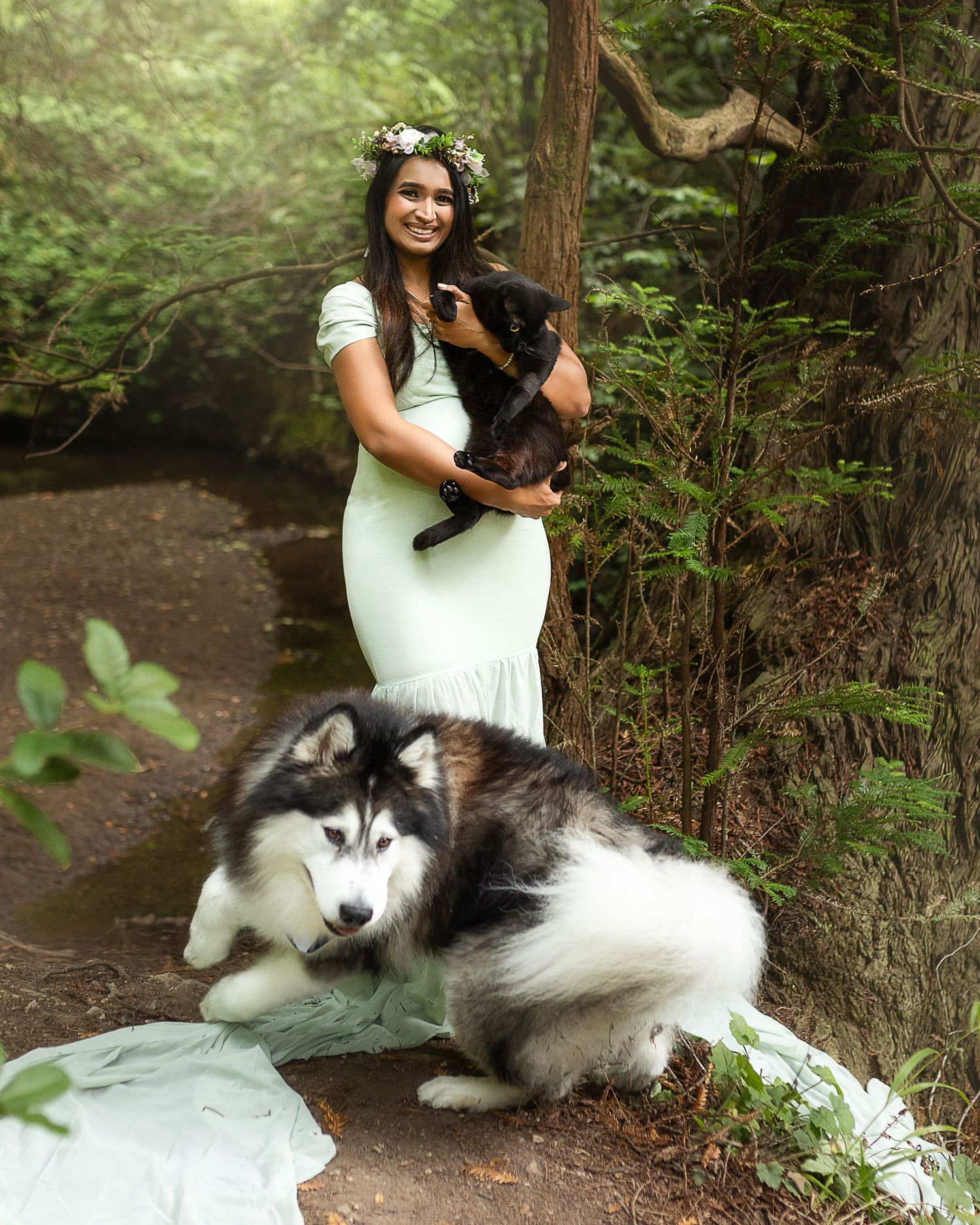 A woman in a mint dress and floral crown holds a black cat while a Husky dog rests nearby in a forest setting.