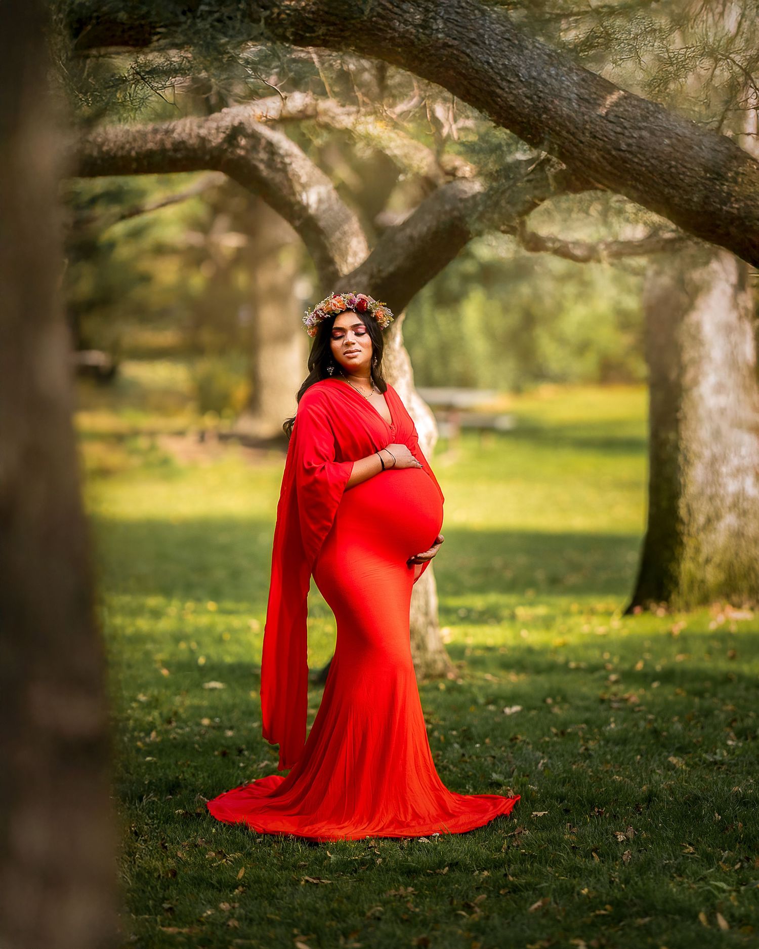 A figure in a flowing red dress stands gracefully among trees in a sunlit outdoor setting during a photo session.