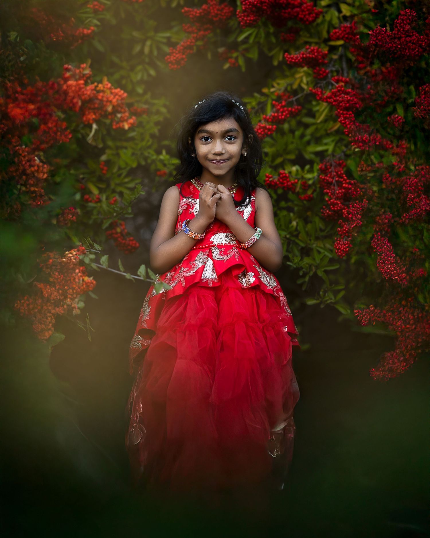 A figure in a vibrant red tulle dress poses gracefully among flowering red bushes in a dreamy outdoor setting.