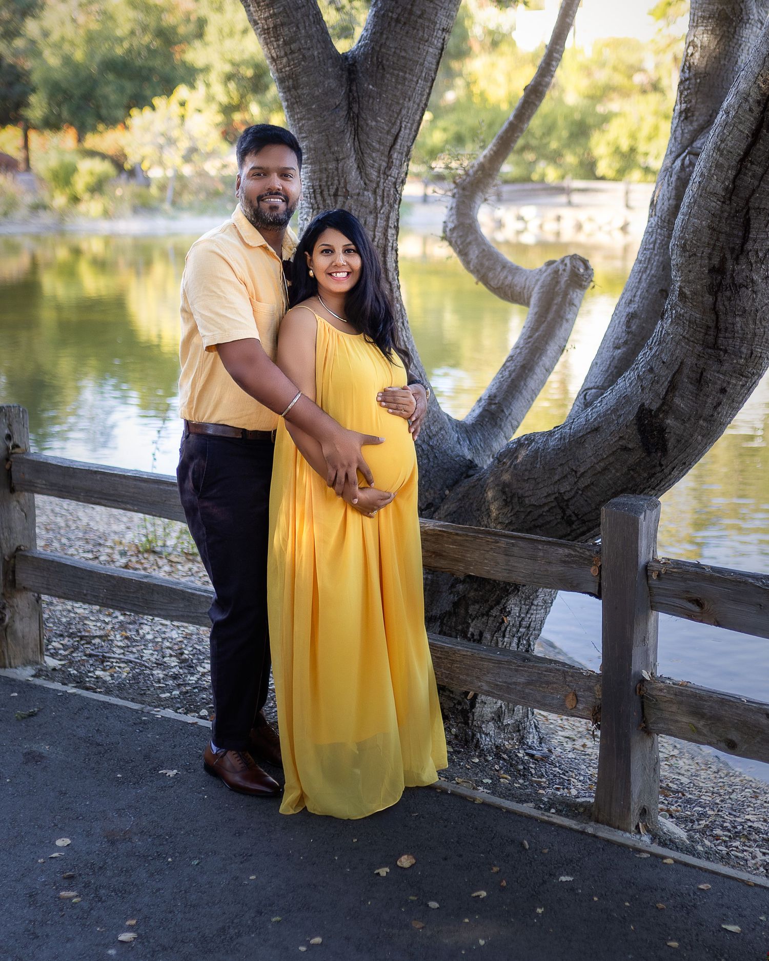 A couple poses together by a lake with a yellow maternity dress and large tree creating a romantic scene.