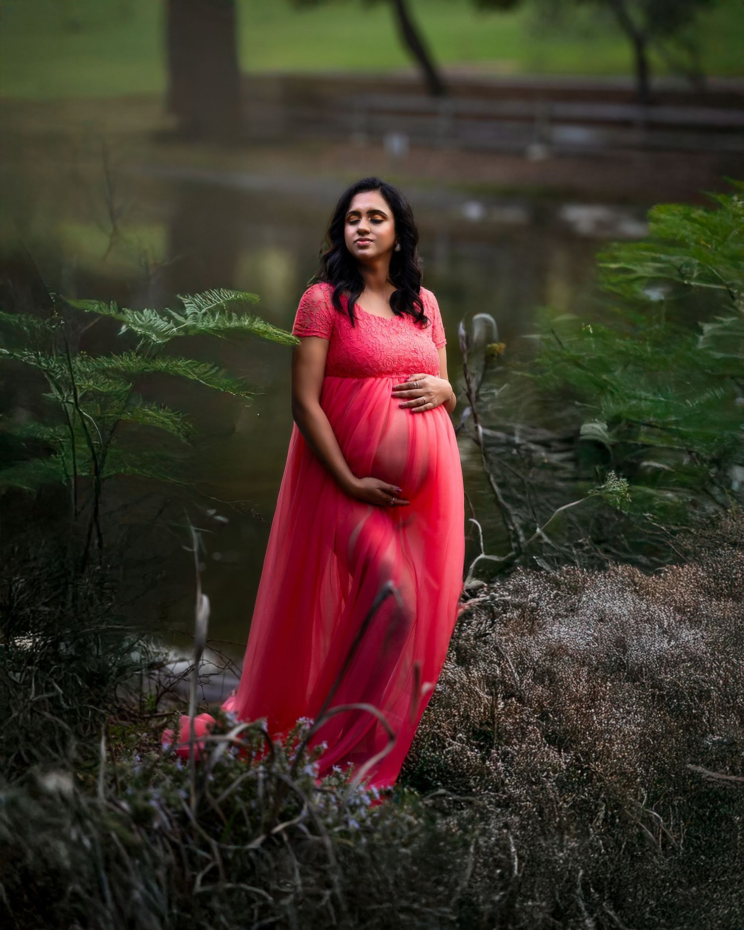 A person in a flowing red gown poses gracefully in a lush garden setting with ferns and greenery.