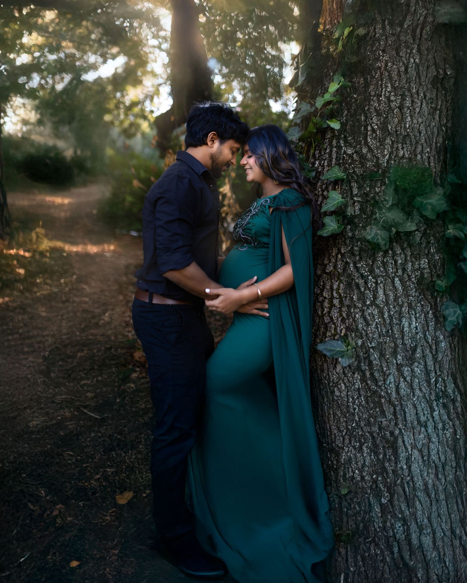 A romantic maternity photo of a couple in a forest setting with sunlit trees, the woman wearing a flowing teal gown.
