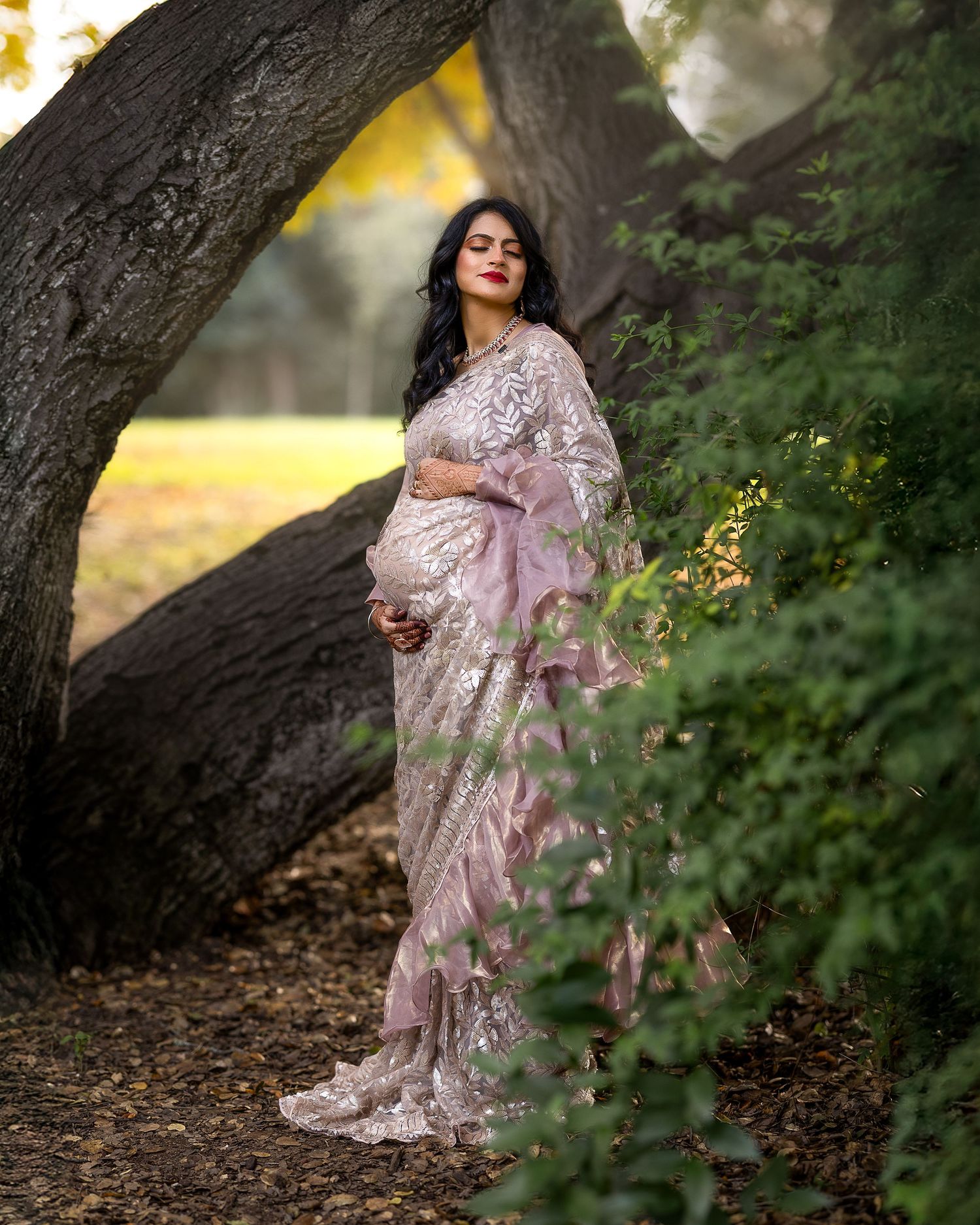 A person in a delicate pink embellished sari poses gracefully against a large tree trunk in a dreamy outdoor setting.