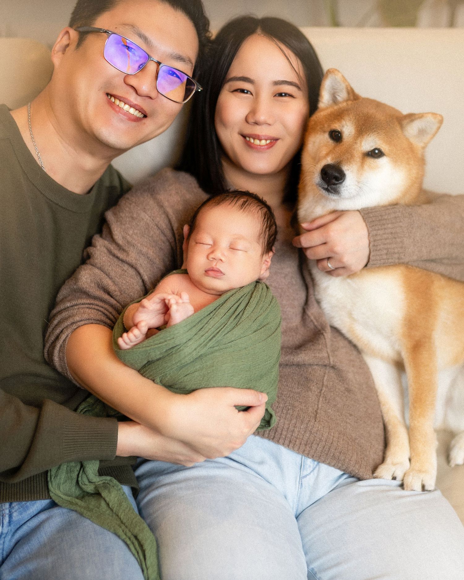 Happy Asian family with newborn baby wrapped in green swaddle and Shiba Inu dog sitting together on couch.