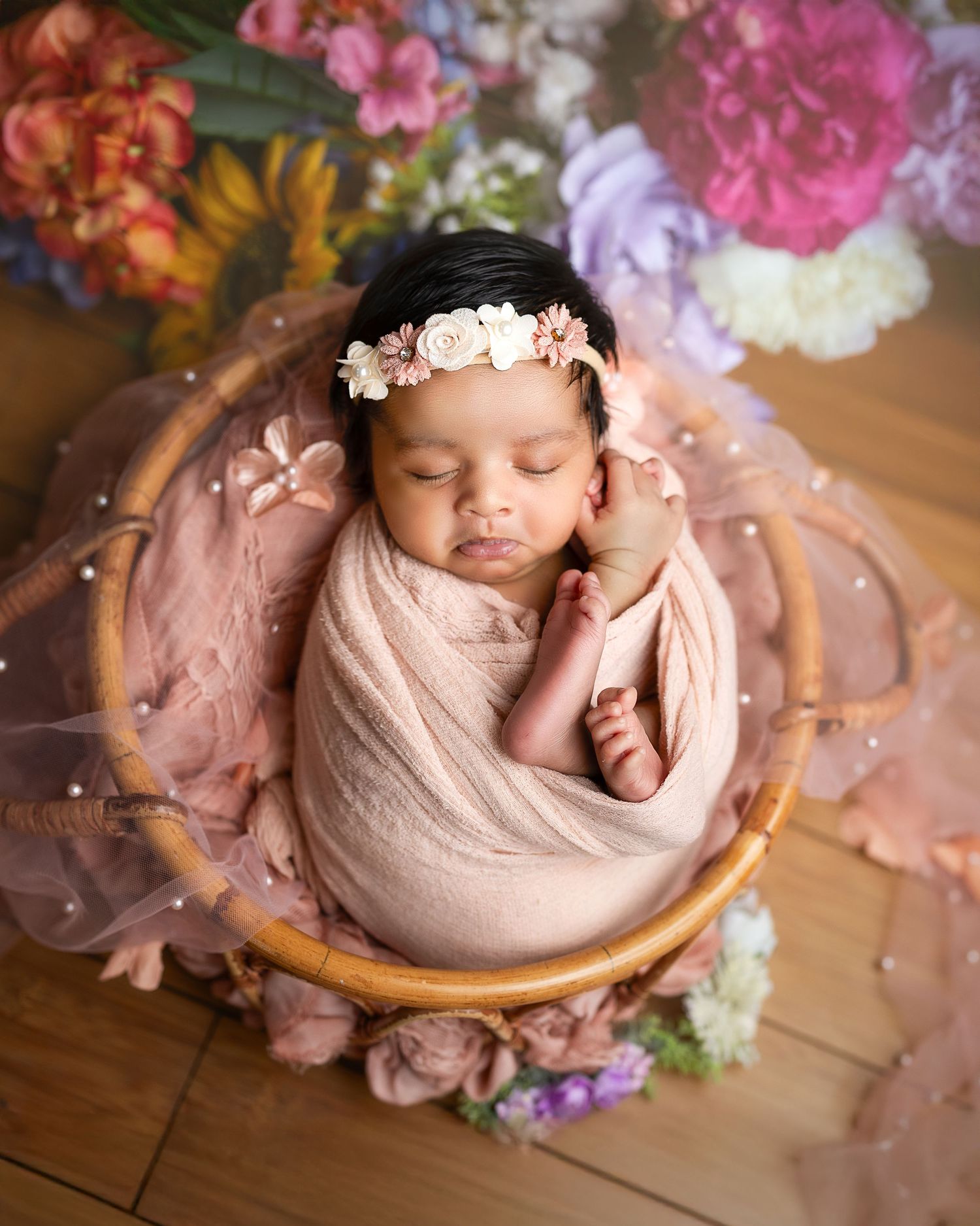 A newborn baby wrapped in pink fabric surrounded by delicate flowers and petals in a rustic wooden setting.