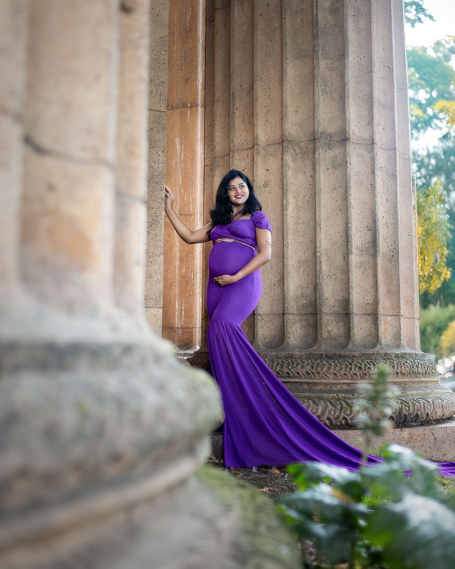 A person in a flowing purple gown poses gracefully between stone columns in an elegant outdoor setting.