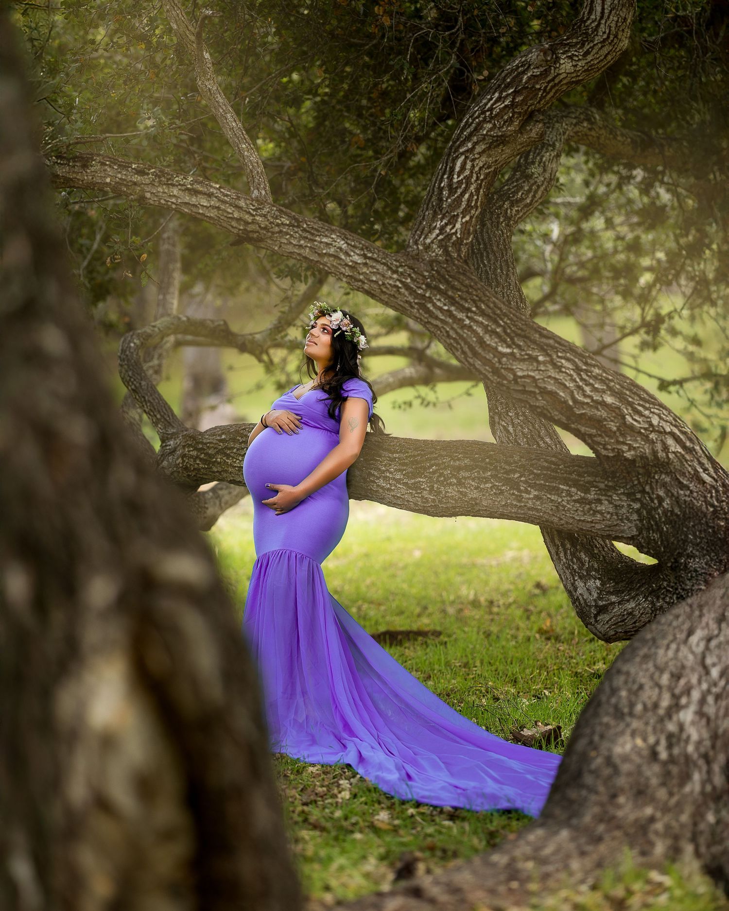 A person in a flowing purple dress poses gracefully on a curved tree branch in a sunlit garden setting.