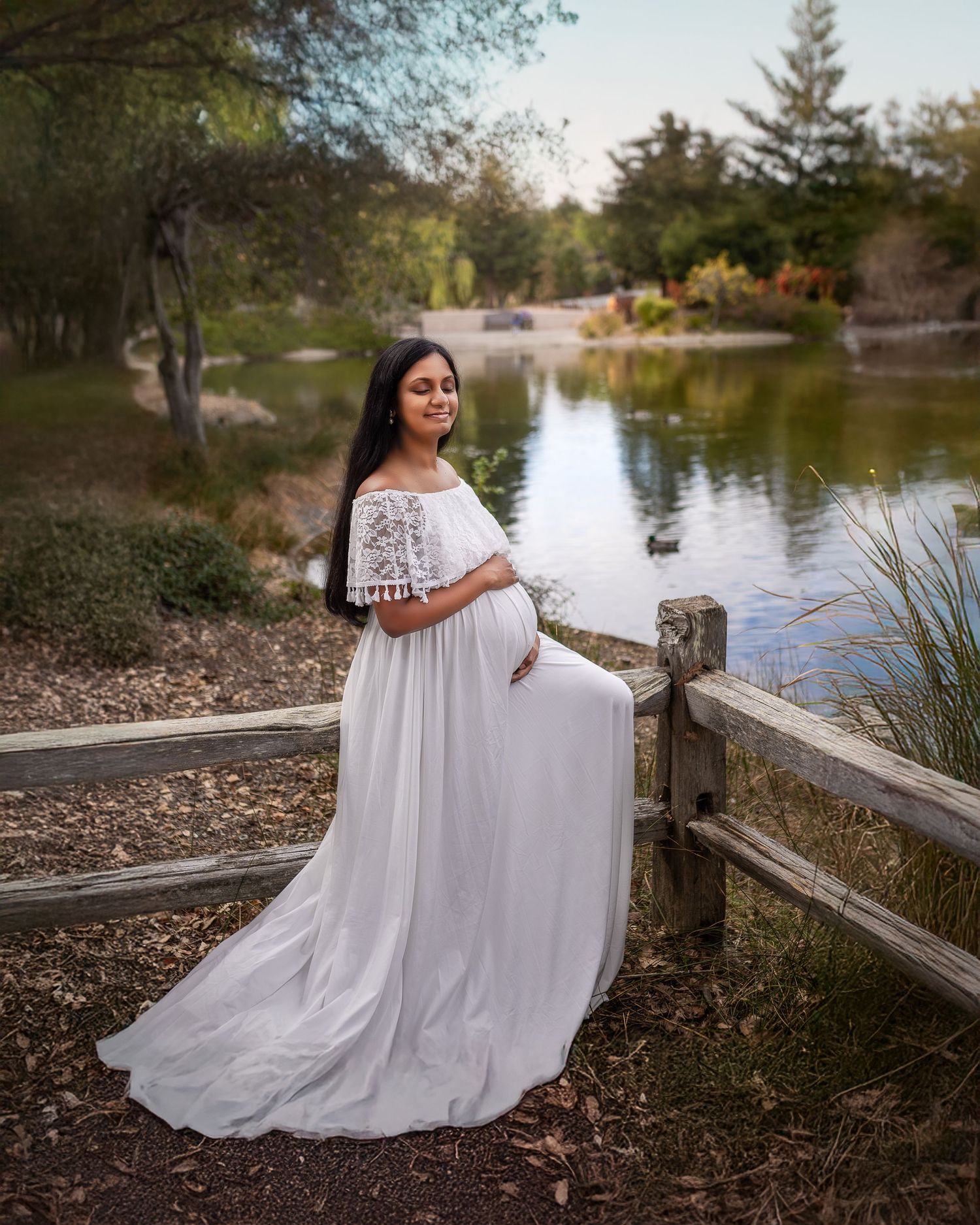 A flowing white dress creates an ethereal silhouette against a tranquil lakeside backdrop during a scenic outdoor photo session.