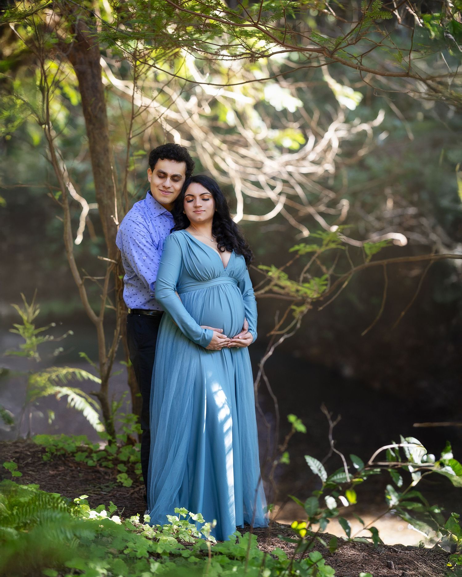 A couple in blue and purple attire embraces in a lush garden setting with dappled sunlight filtering through trees.