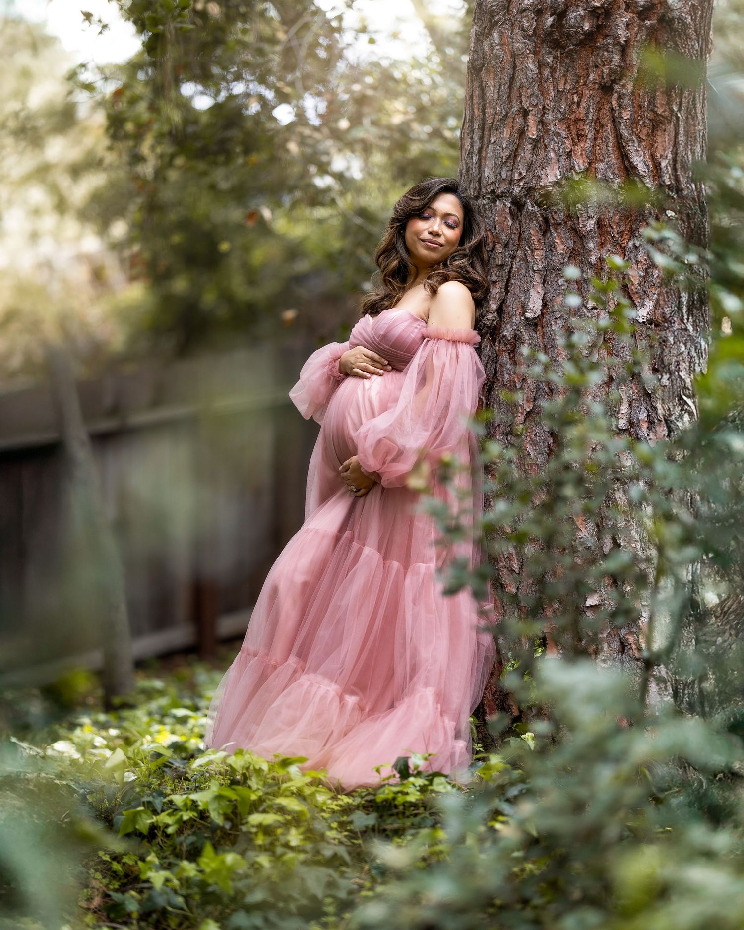 A dreamy portrait session in a garden features a flowing pink tulle gown against lush greenery and tree trunks.