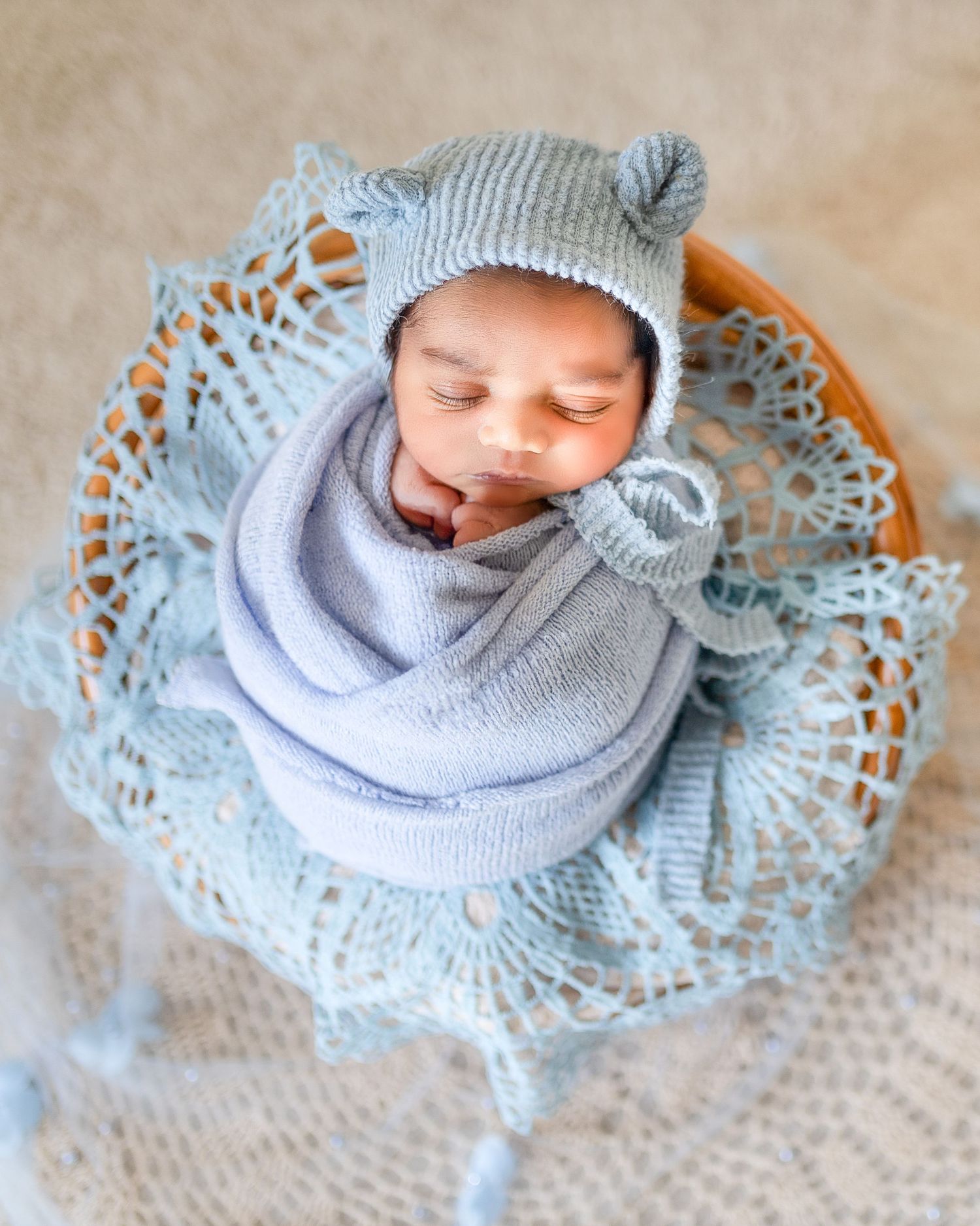 A sleeping baby wearing a gray knit hat and scarf lies on a delicate lace doily against a neutral background.