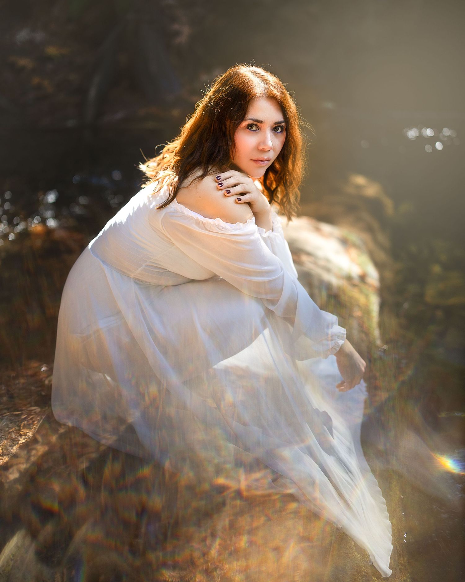 A young woman in a flowing white dress sits gracefully on rocks, bathed in golden sunlight with a dreamy bokeh background.