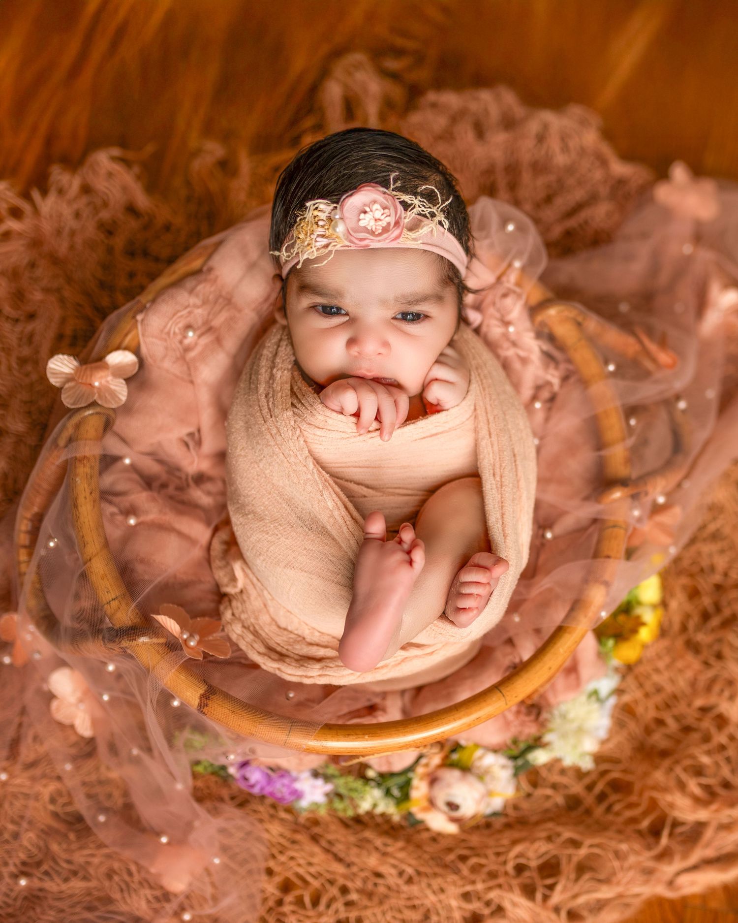 Newborn wearing delicate lace headband rests on peach-colored fabric surrounded by soft floral decorations.