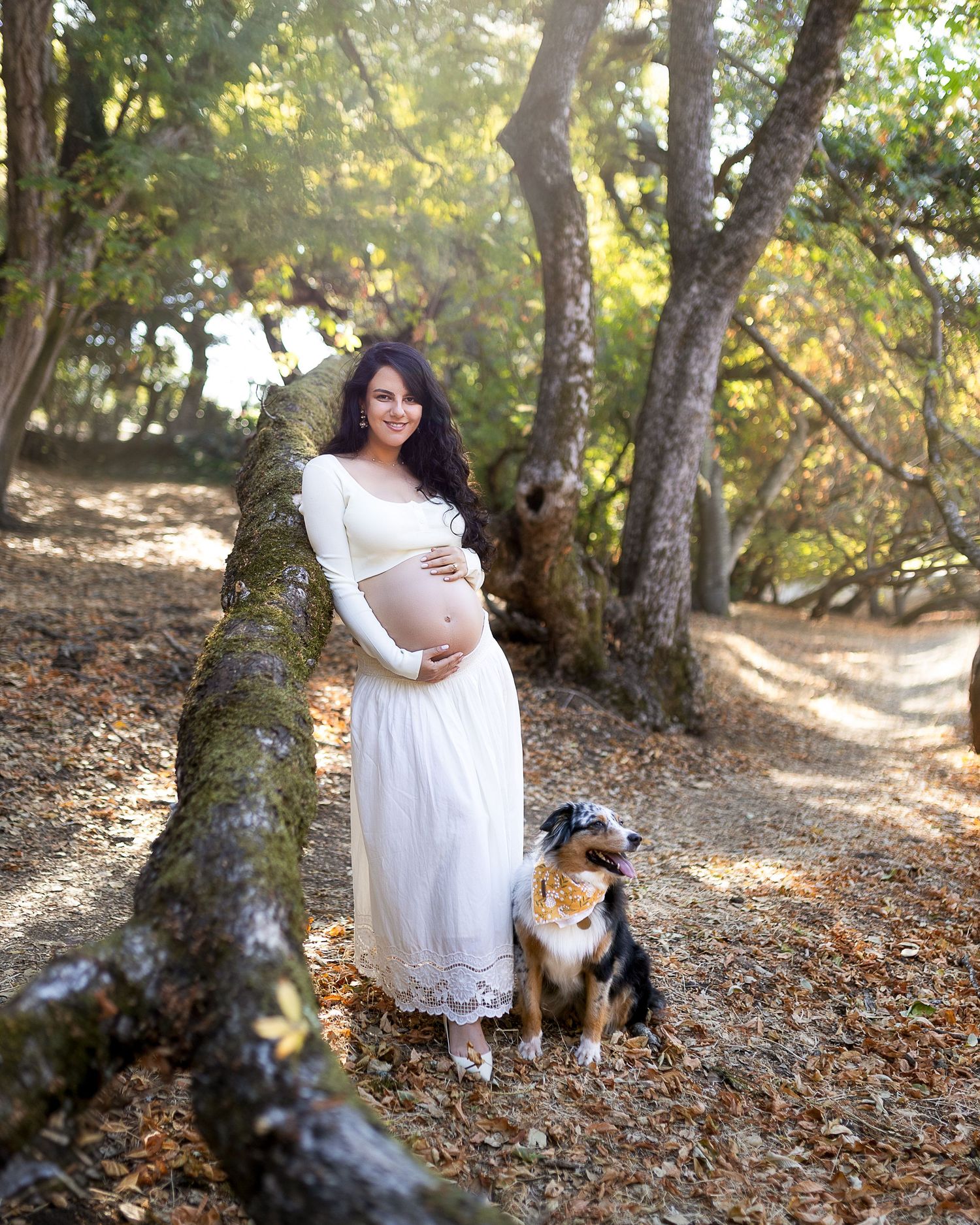 A person in a white dress sits on fallen autumn leaves with two dogs in a wooded outdoor setting.