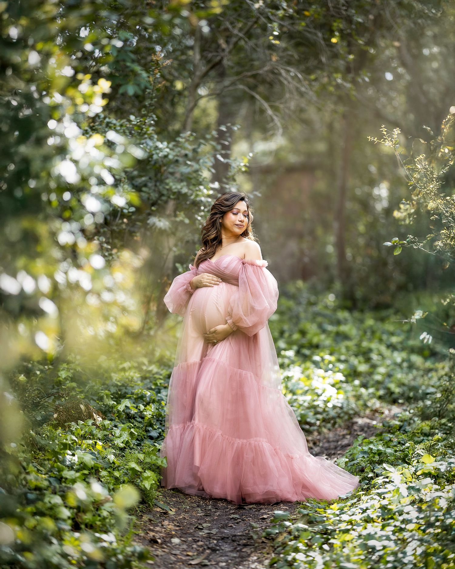 A dreamy maternity portrait in a pink tulle gown surrounded by lush greenery and dappled sunlight in a forest setting.