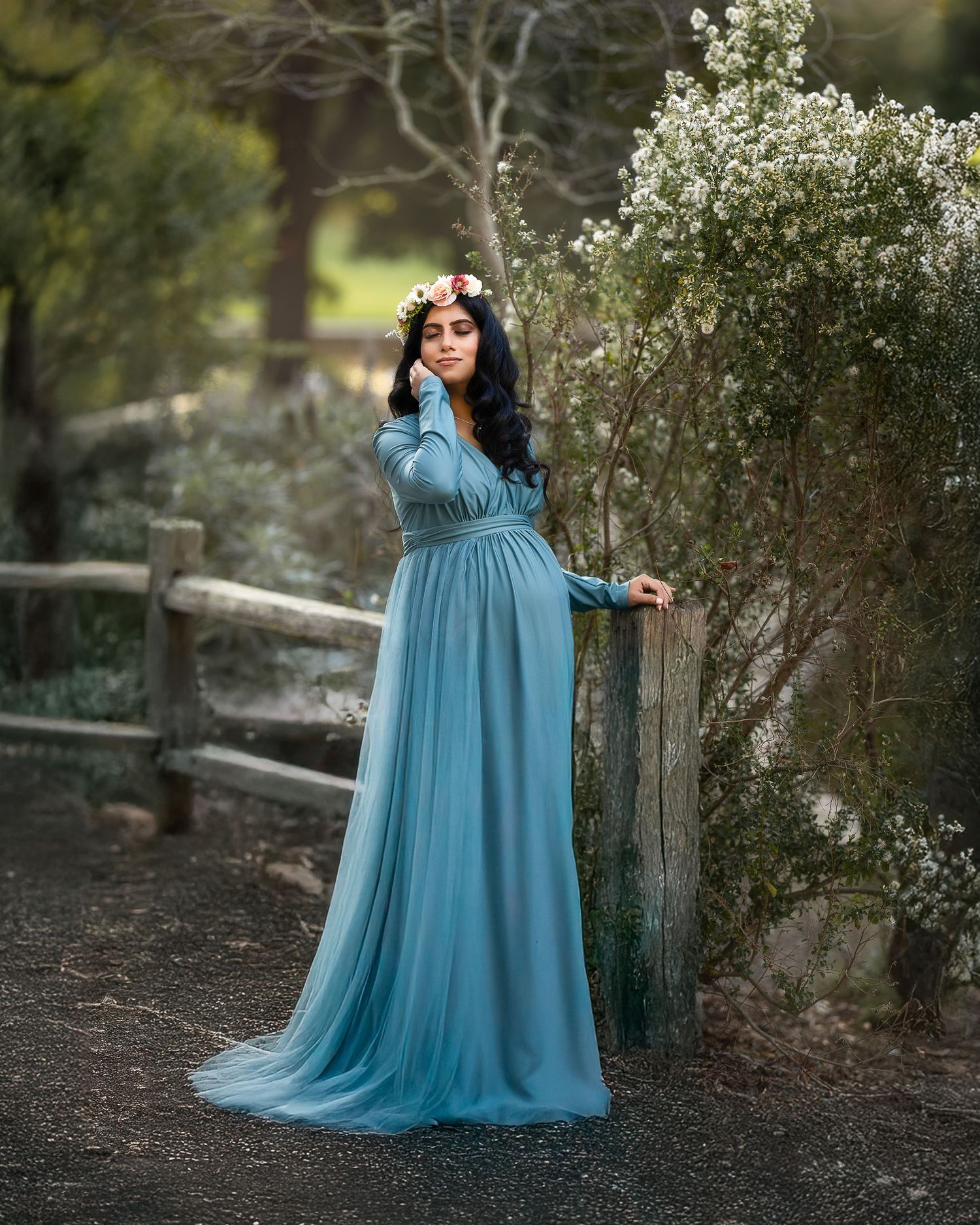 A woman in a flowing turquoise maternity gown poses gracefully in a natural outdoor setting among trees and foliage.