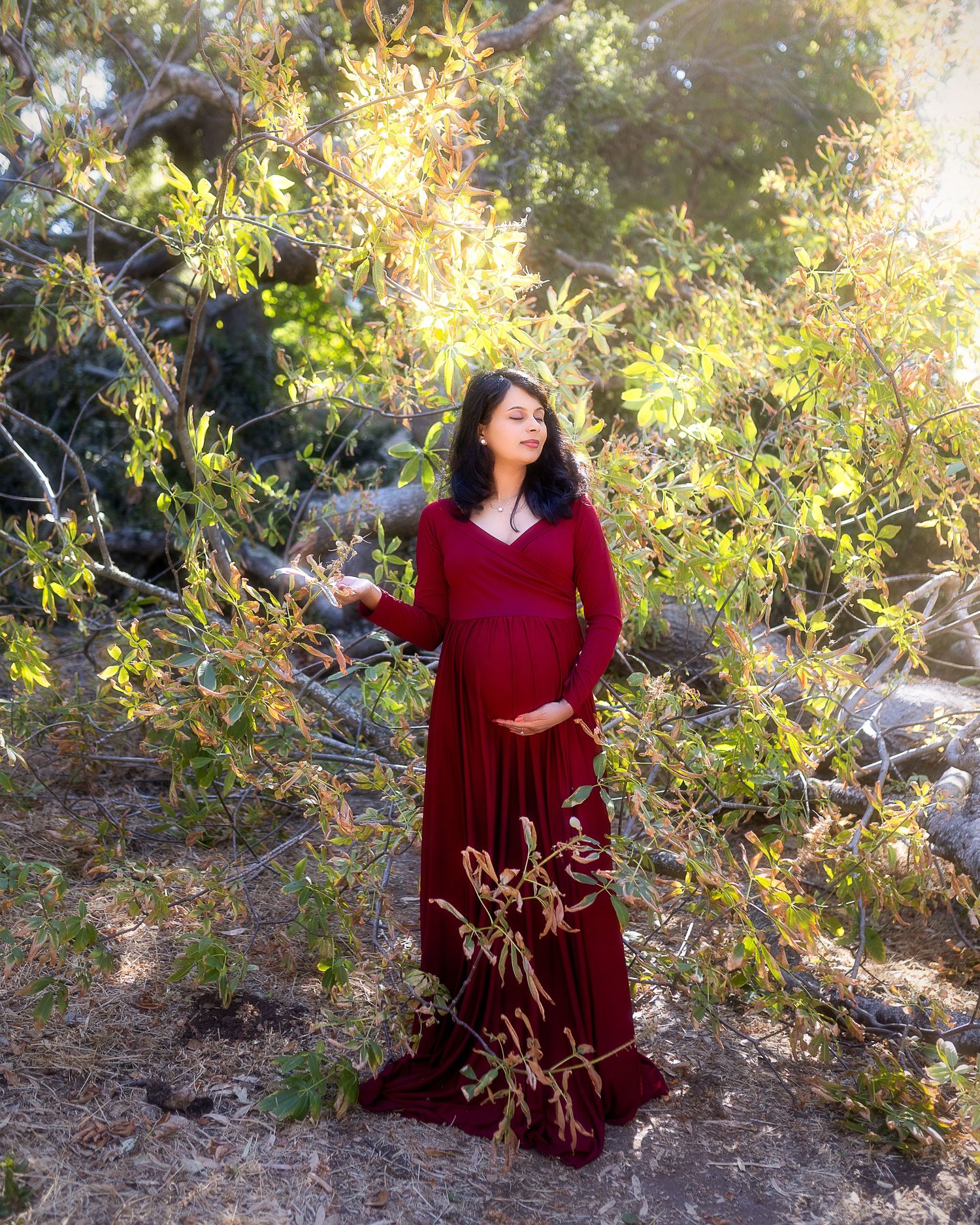 A person in a flowing burgundy velvet dress poses gracefully against a backdrop of sunlit foliage in a natural setting.