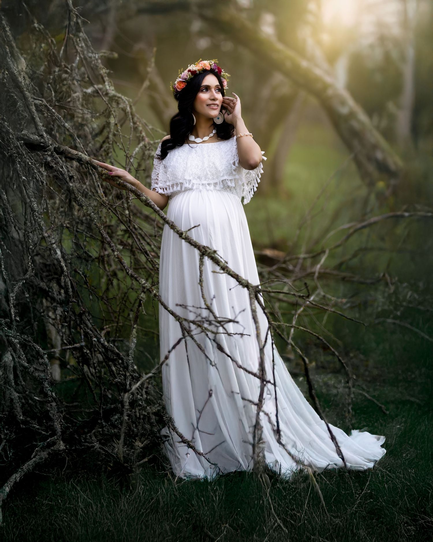 A person in a flowing white dress poses ethereally among dark tree branches in a moody forest setting.