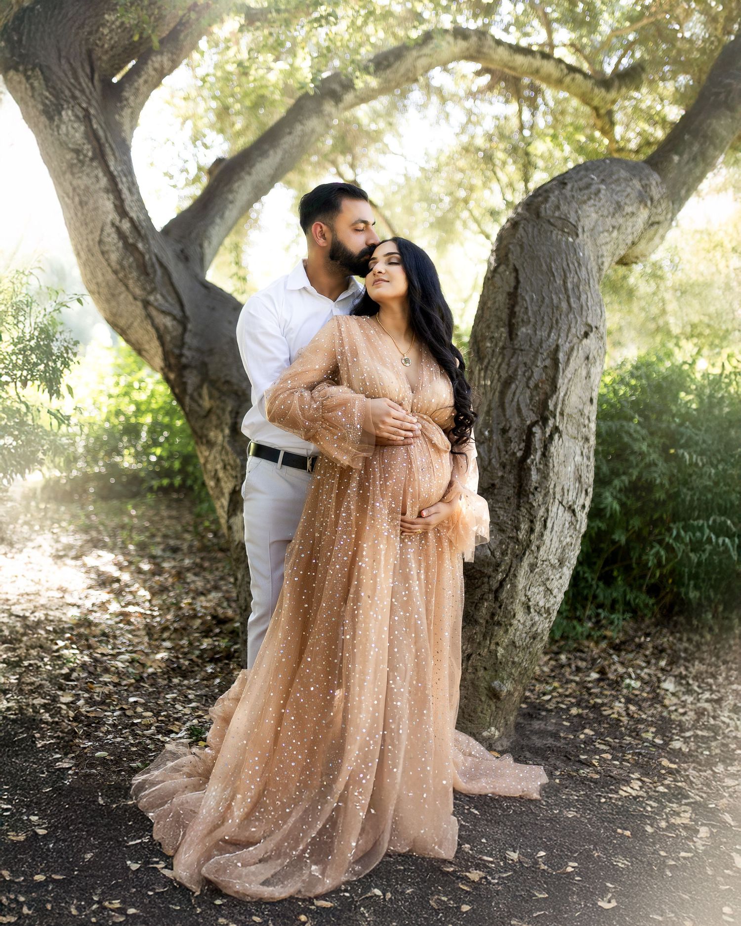A couple poses together under a large tree in a natural setting, with one wearing a flowing peach-colored sparkly gown.