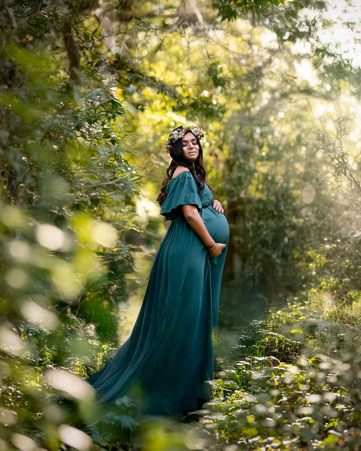A person in a flowing teal gown poses gracefully in a sunlit forest setting with dreamy bokeh effects.